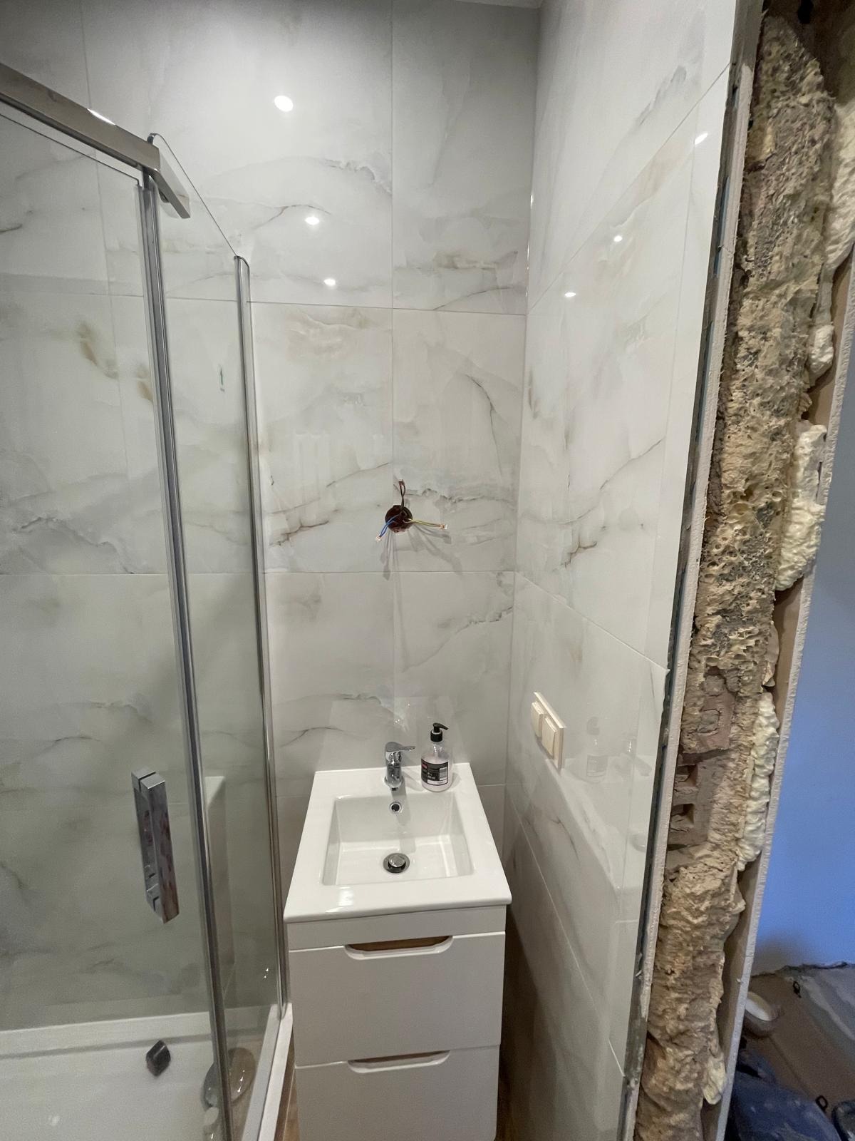 Biała łazienka z kabiną prysznicową i umywalką na szafce. Widoczna ściana w trakcie prac wykończeniowych z pianką montażową. Marmurowe płytki na ścianach.