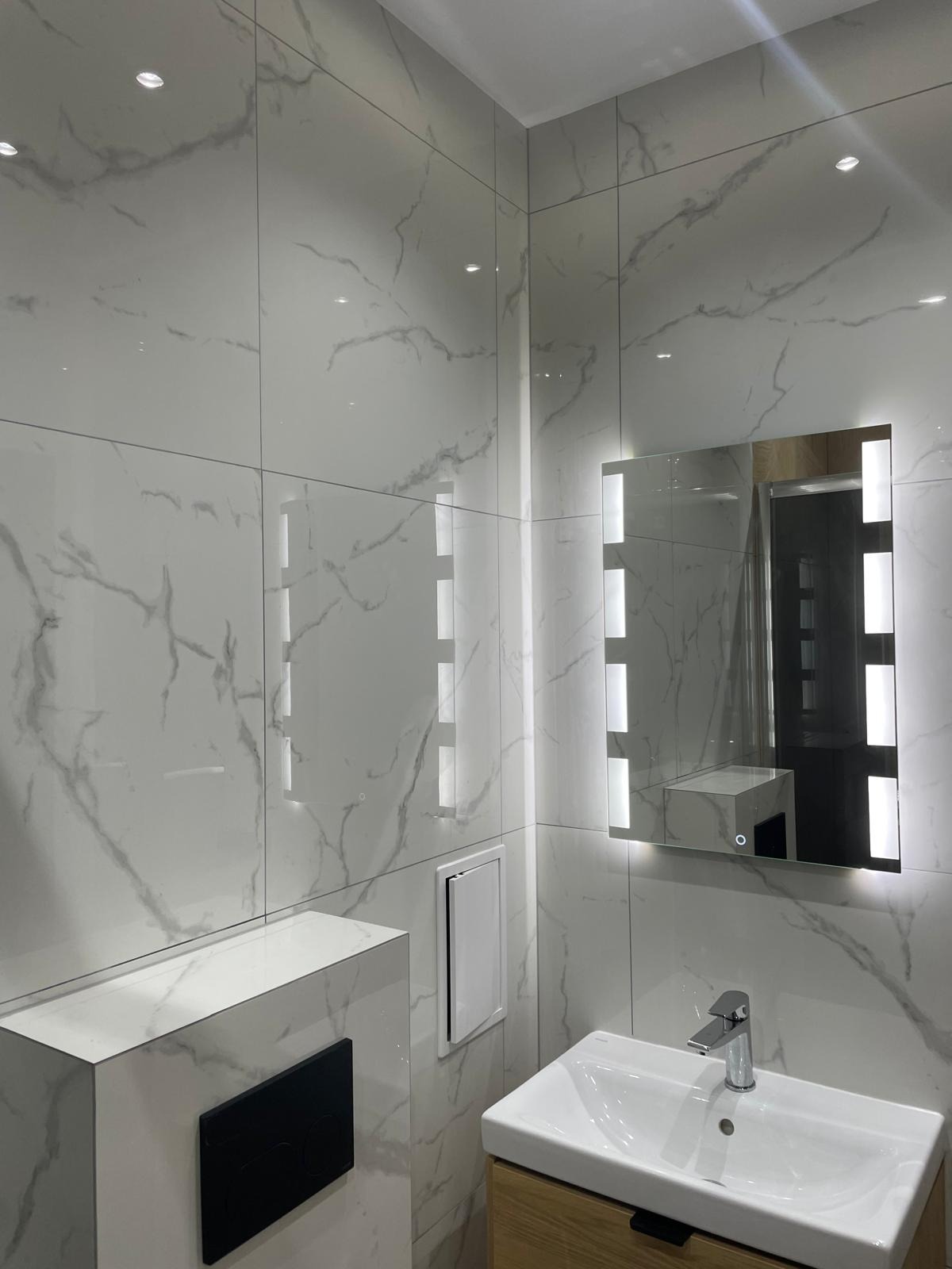 Nowoczesna łazienka z białymi płytkami imitującymi marmur, podświetlanym lustrem i drewnianą szafką pod umywalką. Minimalistyczny design z czarnymi akcentami.