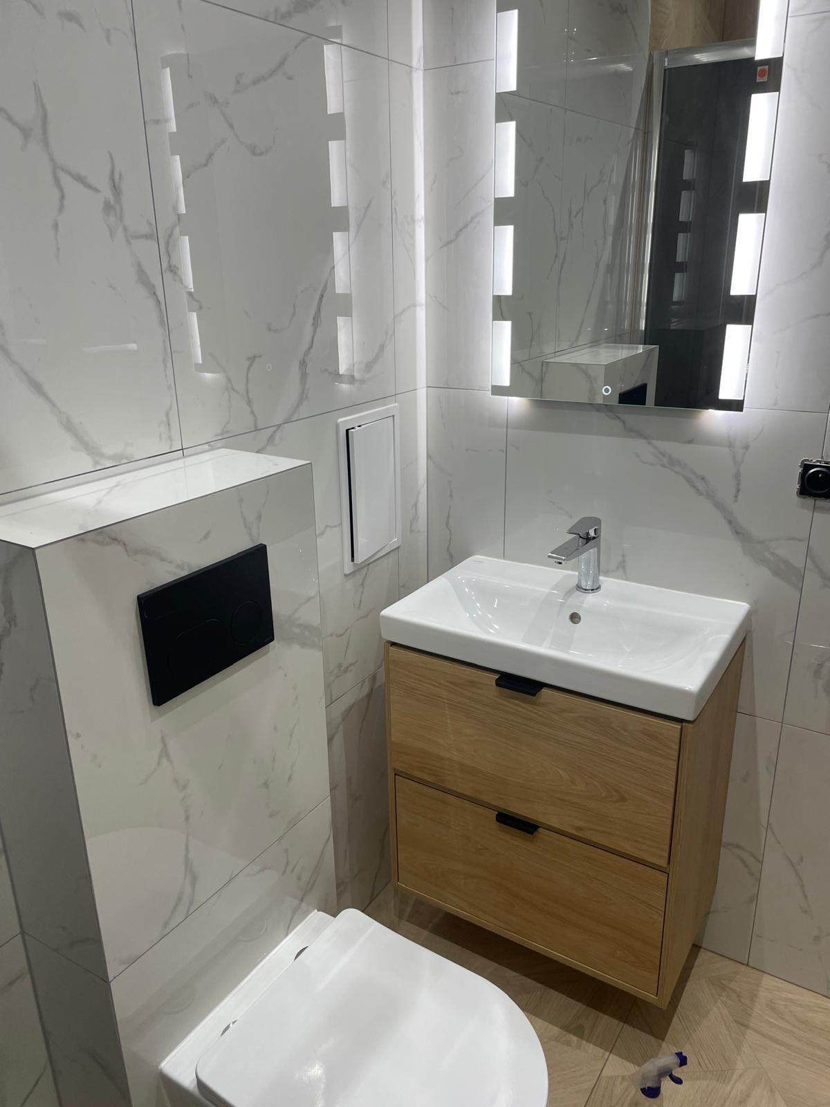 Nowoczesna łazienka z marmurowymi płytkami, podświetlanym lustrem i drewnianą szafką pod umywalką. Widoczny fragment toalety i czarny przycisk spłuczki.