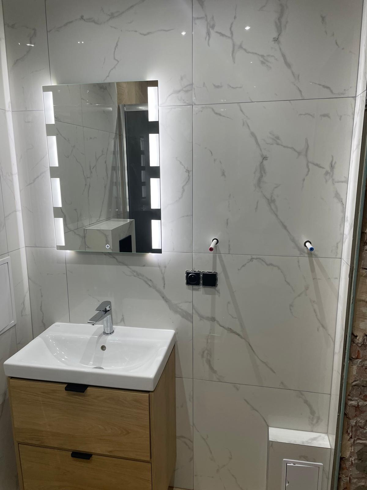 Wykończona łazienka z umywalką na szafce z drewnianym fornirem, lustrem z podświetleniem LED i białymi płytkami imitującymi marmur. Widoczne gniazdka i instalacja wodna.