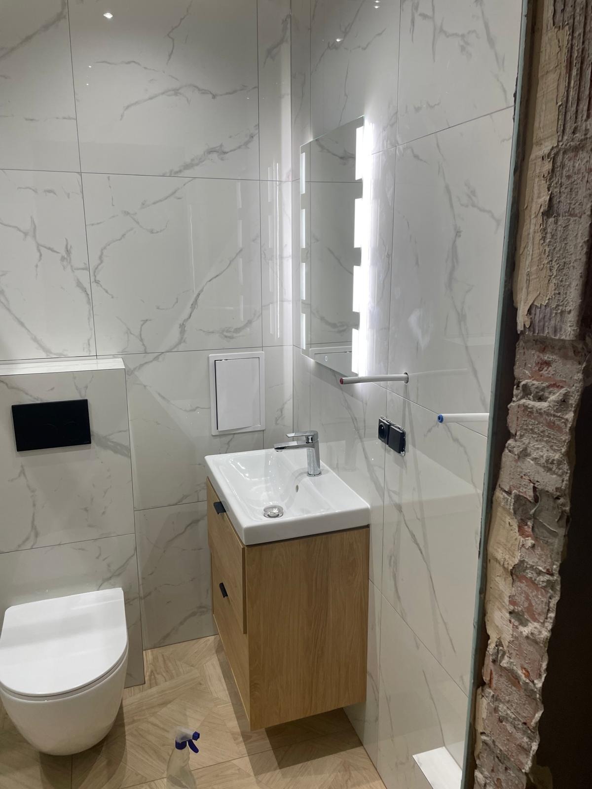 Nowoczesna łazienka po remoncie: białe płytki imitujące marmur, drewniana szafka pod umywalką, podświetlane lustro. Fragment ściany w stanie surowym po prawej.