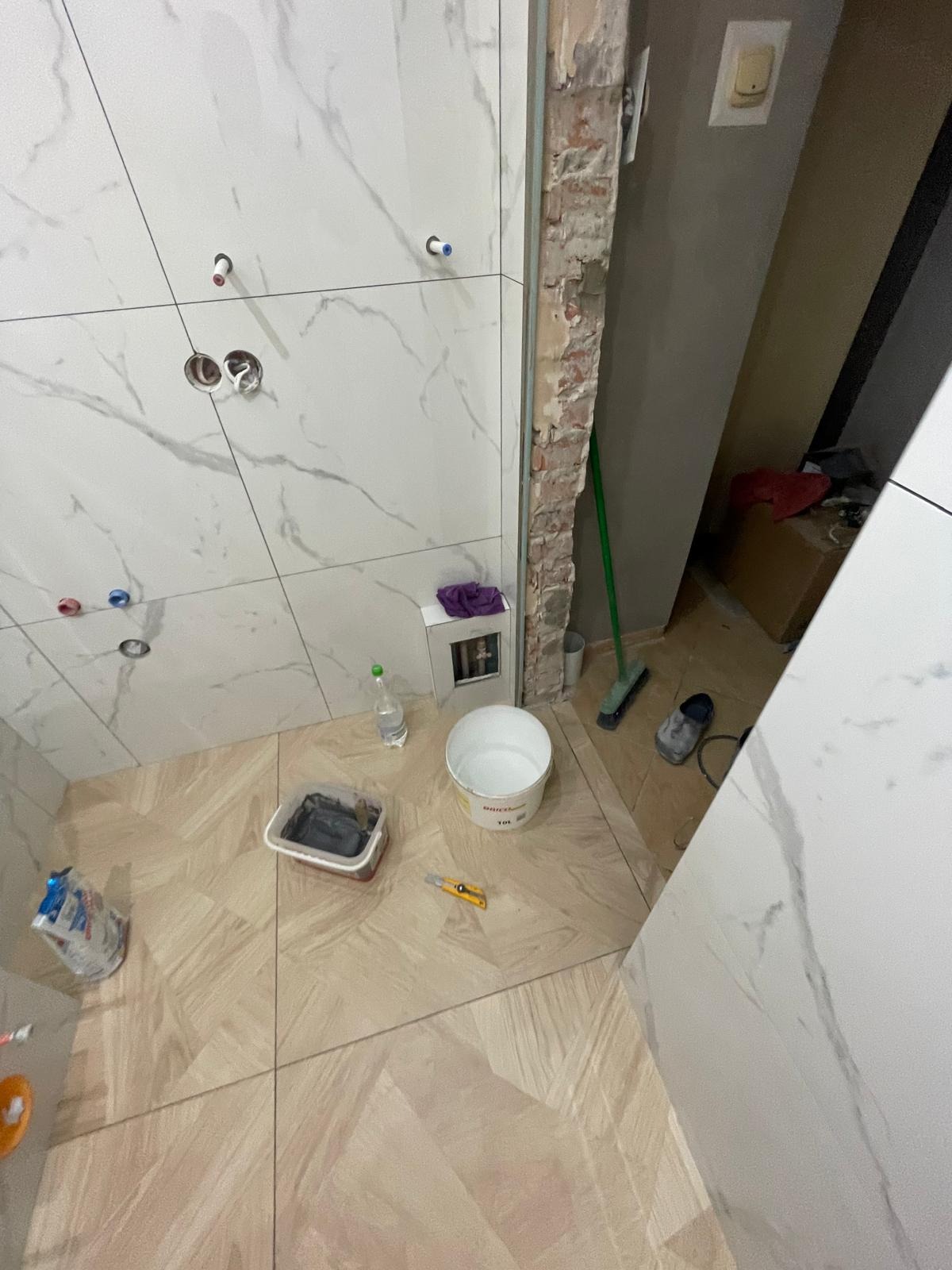 Łazienka w trakcie prac: białe płytki imitujące marmur na ścianach, beżowe płytki na podłodze. Widoczne narzędzia i materiały budowlane. Otwór w ścianie z odsłoniętą cegłą.