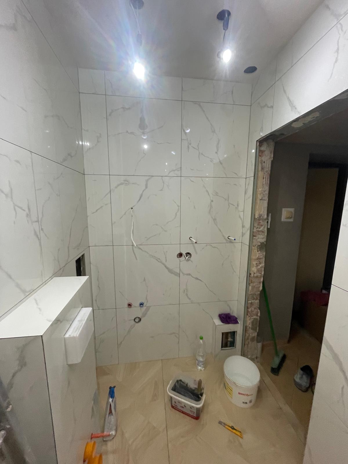 Biała łazienka z marmurowymi płytkami w trakcie wykańczania. Widoczne narzędzia glazurnicze i instalacja. Otwór drzwiowy bez ościeżnicy.