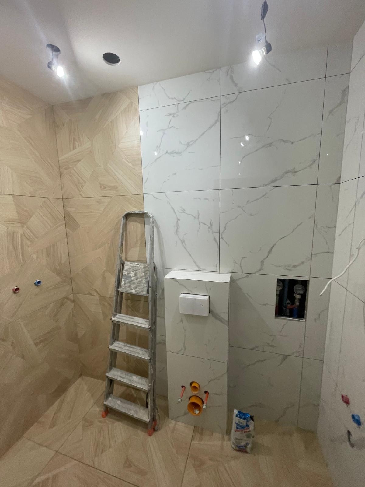 Łazienka w trakcie wykończenia: częściowo ułożone płytki imitujące marmur i drewno, drabina malarska, instalacja podtynkowa WC, widoczne rury.