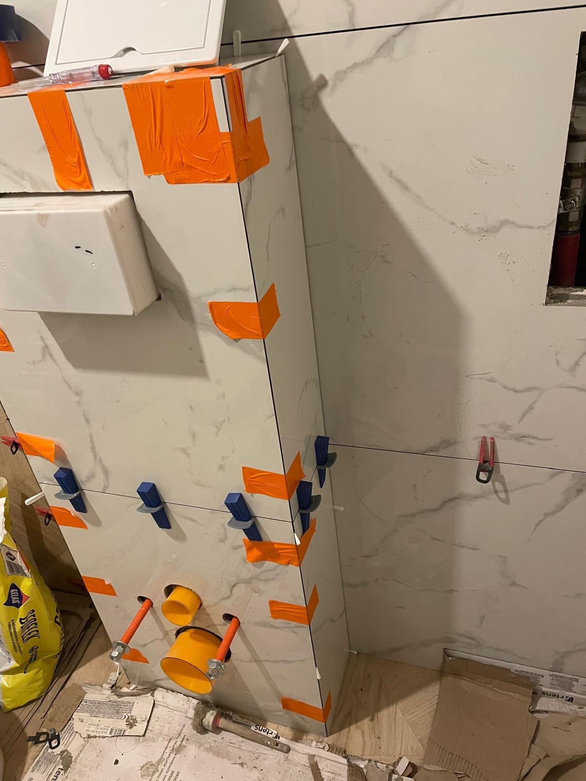 Montaż płytek imitujących marmur w łazience, z użyciem pomarańczowej taśmy i niebieskich klipsów poziomujących. Widoczne rury i elementy instalacji.