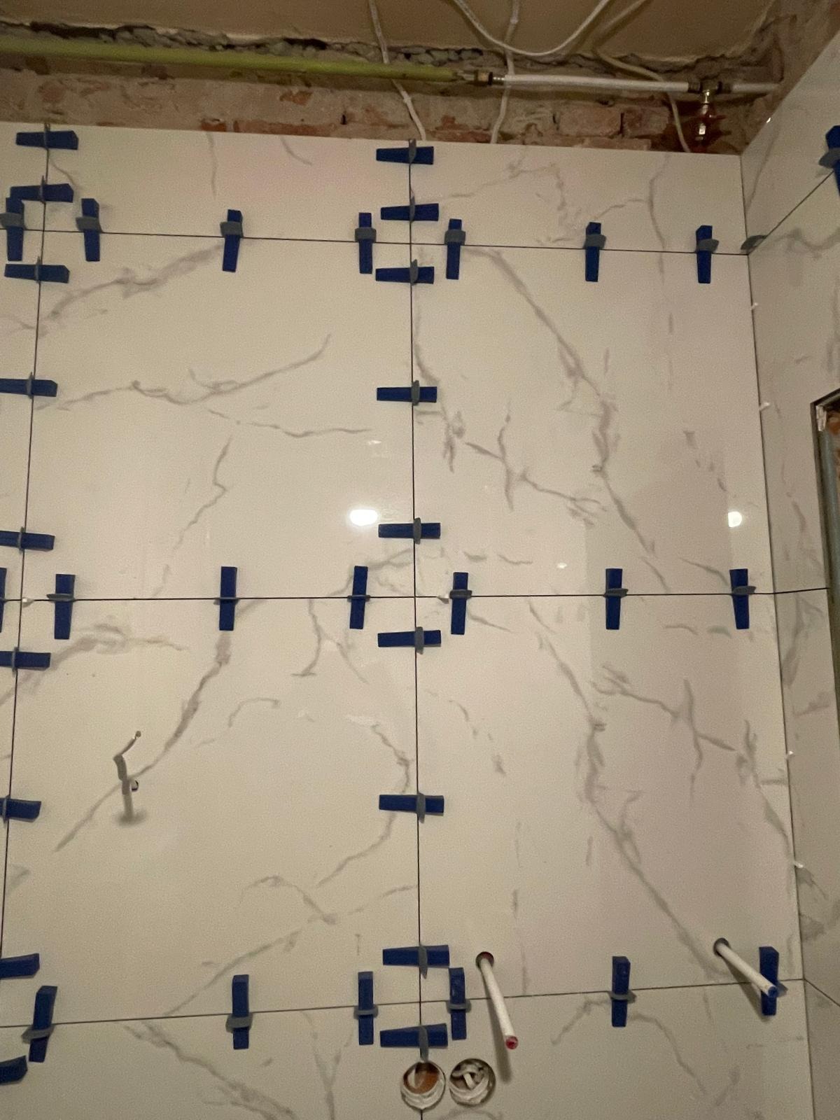 Ściana z białych płytek imitujących marmur, z niebieskimi klinami i sznurkiem do poziomowania, widoczne rury i niedokończone wykończenie u góry.