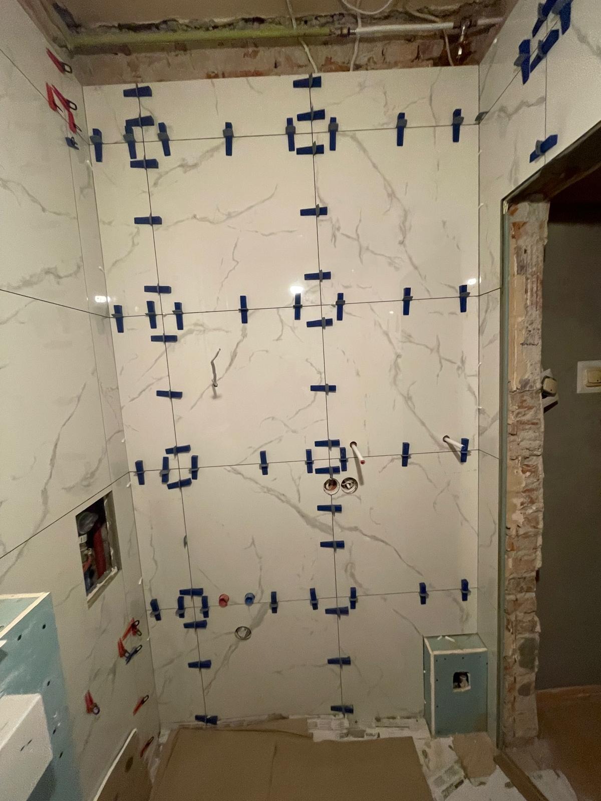 Ściana w łazience z białymi płytkami imitującymi marmur, zabezpieczonymi niebieskimi krzyżykami dystansowymi i żyłką, widoczne instalacje i fragment wejścia.
