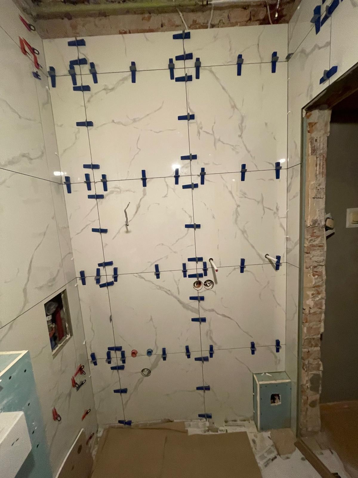 Ściana łazienki w trakcie układania płytek imitujących marmur, z użyciem systemu poziomowania (klipsy i kliny). Widoczne przewody instalacji wodnej i elektrycznej.