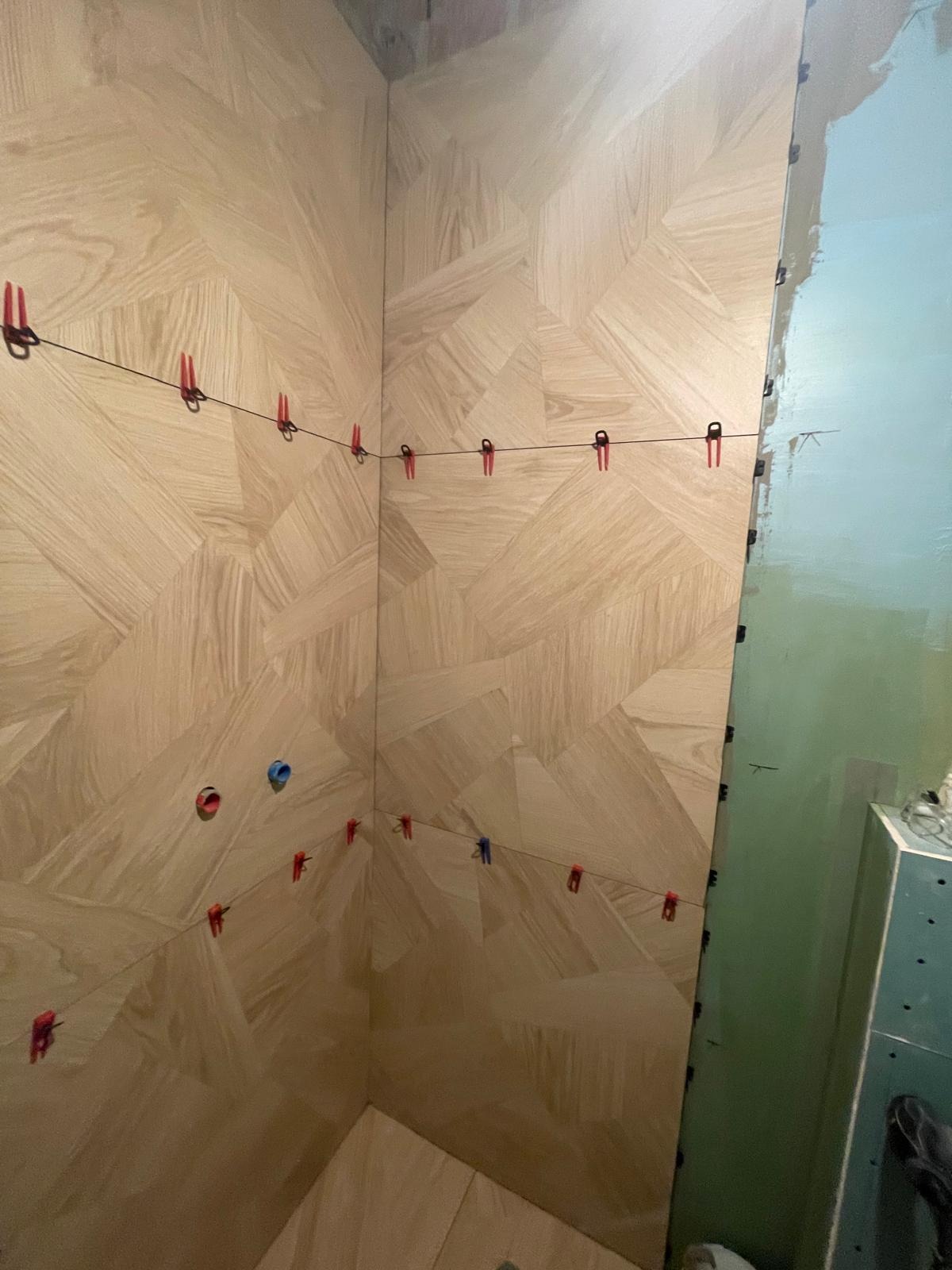 Układanie płytek imitujących drewno w rogu łazienki, z użyciem klipsów poziomujących. Widoczne świeżo położone płytki i przygotowana ściana do dalszych prac.