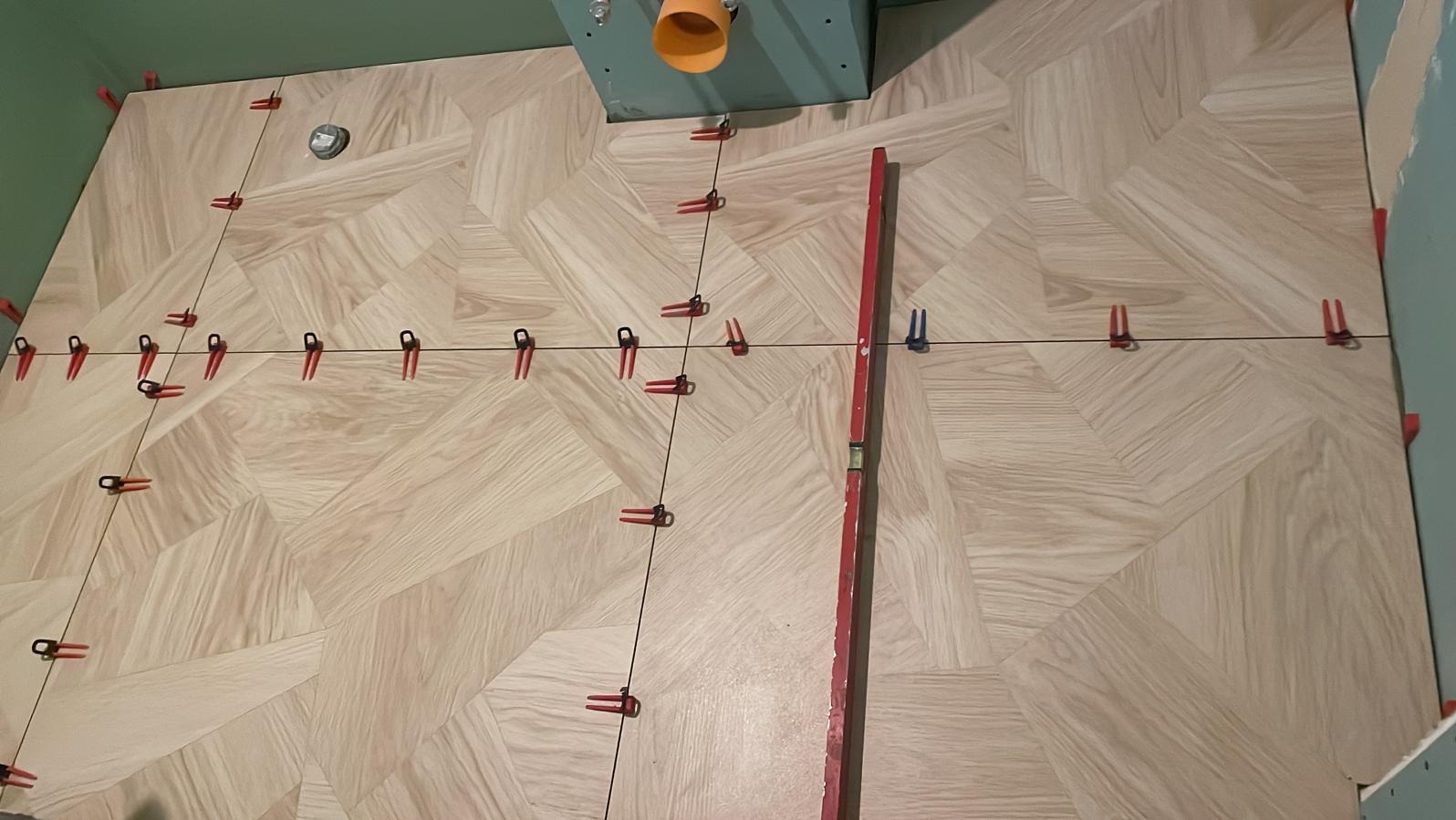 Ułożone płytki podłogowe w jodełkę, z czerwonymi klipsami poziomującymi i sznurkiem, widok z góry. Częściowo widoczny poziomica i fragment instalacji.
