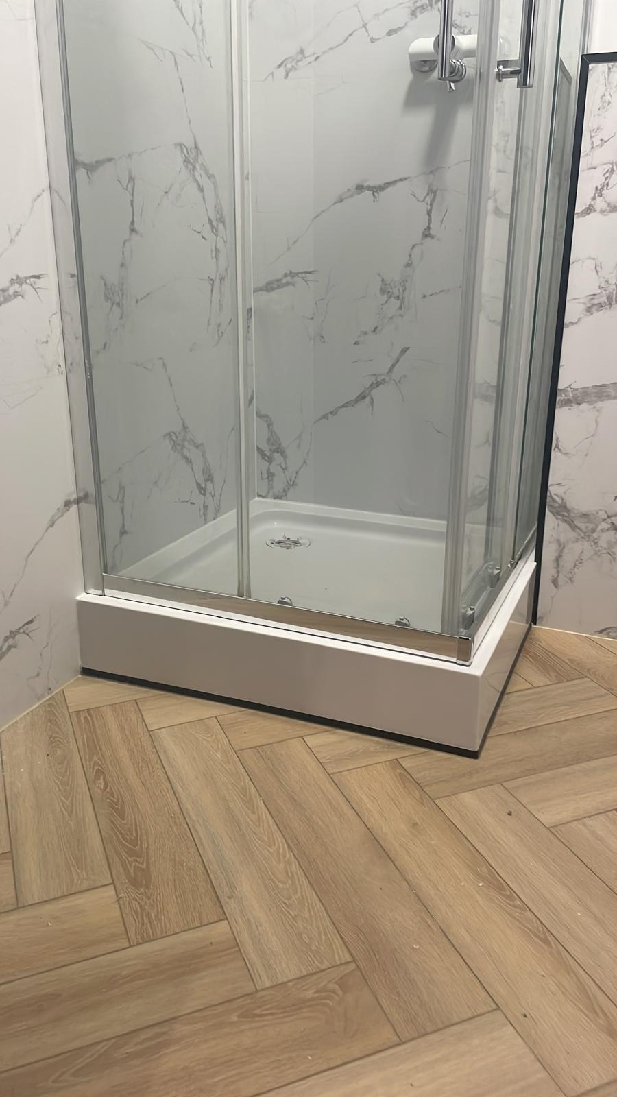 Nowoczesna kabina prysznicowa z białym brodzikiem i marmurowymi płytkami ściennymi, uzupełniona podłogą z paneli ułożonych w jodełkę. Minimalistyczny design.