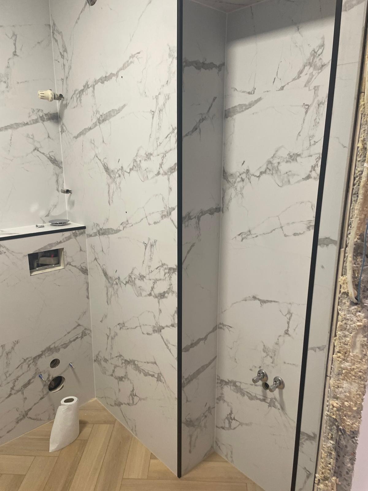 Łazienka w trakcie wykańczania: białe płytki imitujące marmur z czarnymi listwami, drewnopodobna podłoga, widoczne instalacje i fragment ściany w stanie surowym.