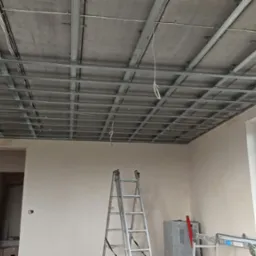 Stelaż metalowy podwieszanego sufitu zamontowany w surowym wnętrzu, widoczna drabina aluminiowa i okno PCV