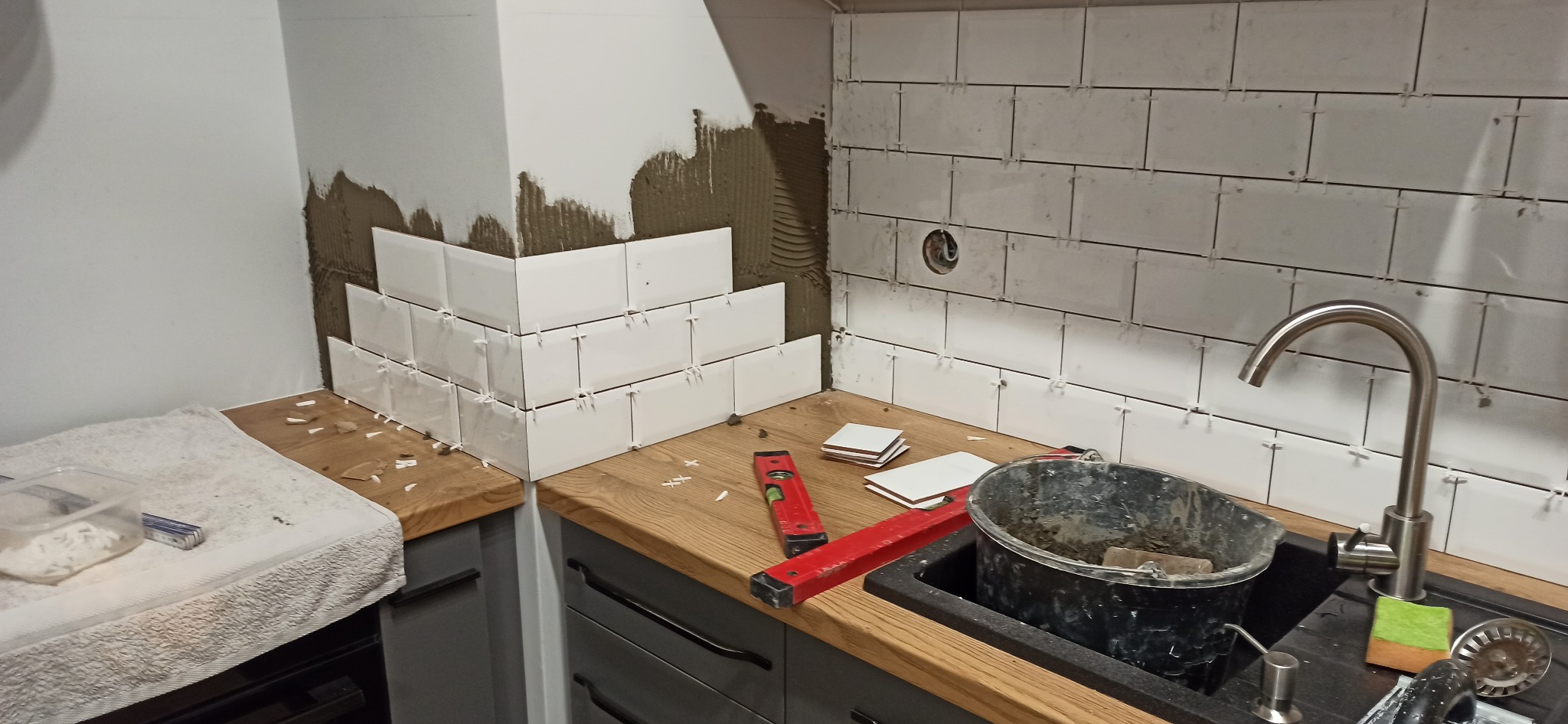 Układanie białych, prostokątnych płytek na ścianie w kuchni, widoczne poziomice, wiadro z zaprawą i elementy instalacji wodnej