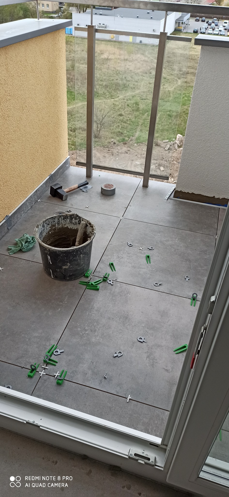 Układanie szarych płytek na balkonie z widokiem na zewnątrz, widoczny kubeł z zaprawą, młotek, taśma klejąca i plastikowe krzyżyki dystansowe.