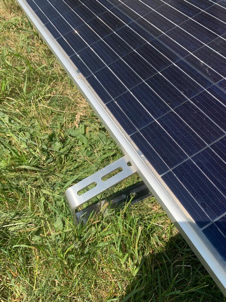 Fragment panelu fotowoltaicznego z aluminiowym uchwytem montażowym FastLine Garden na tle trawy. Widoczne dwa otwory w uchwycie.