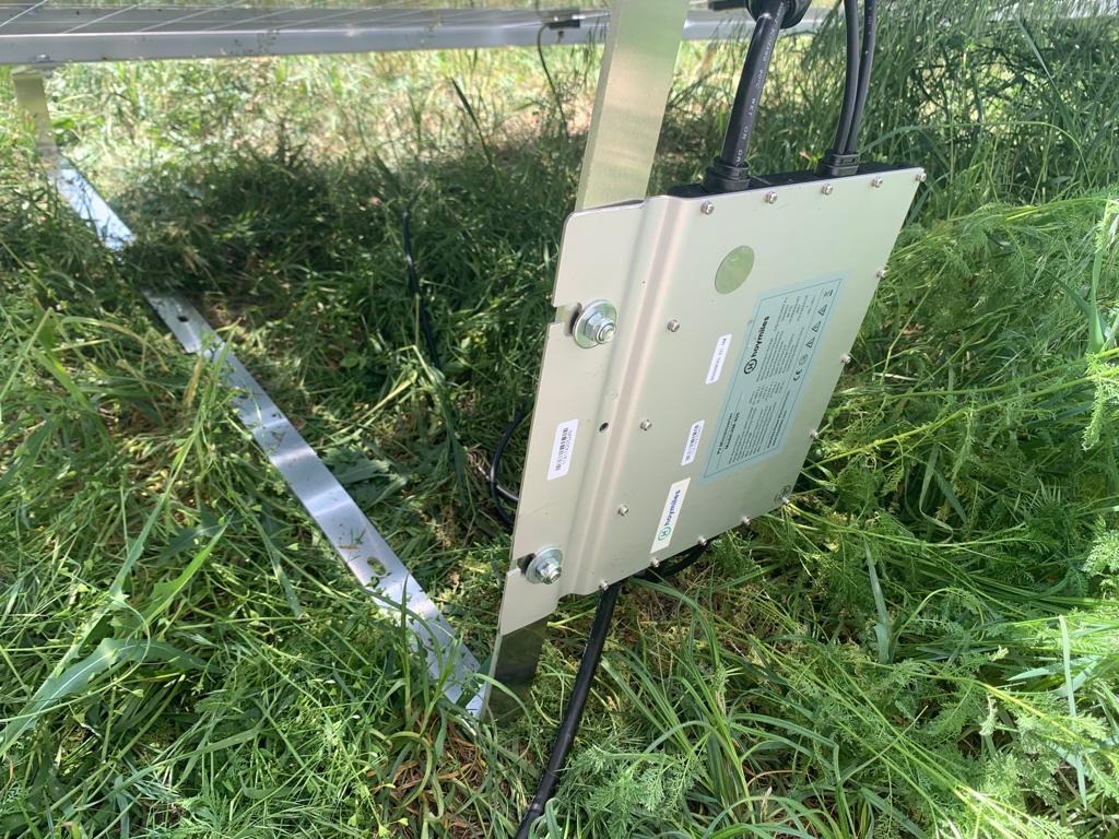 Mikroinwerter Hoymiles zamontowany pod panelem słonecznym, widoczne kable i konstrukcja montażowa, całość umieszczona w trawie.