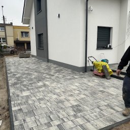 Magnolia zakładanie ogrodów - Ułożona szara kostka brukowa przed nowoczesnym domem. Widoczna ubijarka do kostki i fragment osoby. Kostka ułożona w nieregularny wzór.