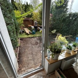 Magnolia zakładanie ogrodów - Przygotowanie terenu pod trawnik z rolki w Mosinie. Widok przez okno na wyrównaną ziemię, narzędzia i deski. Prace ogrodowe w toku, zielona roślinność w tle.