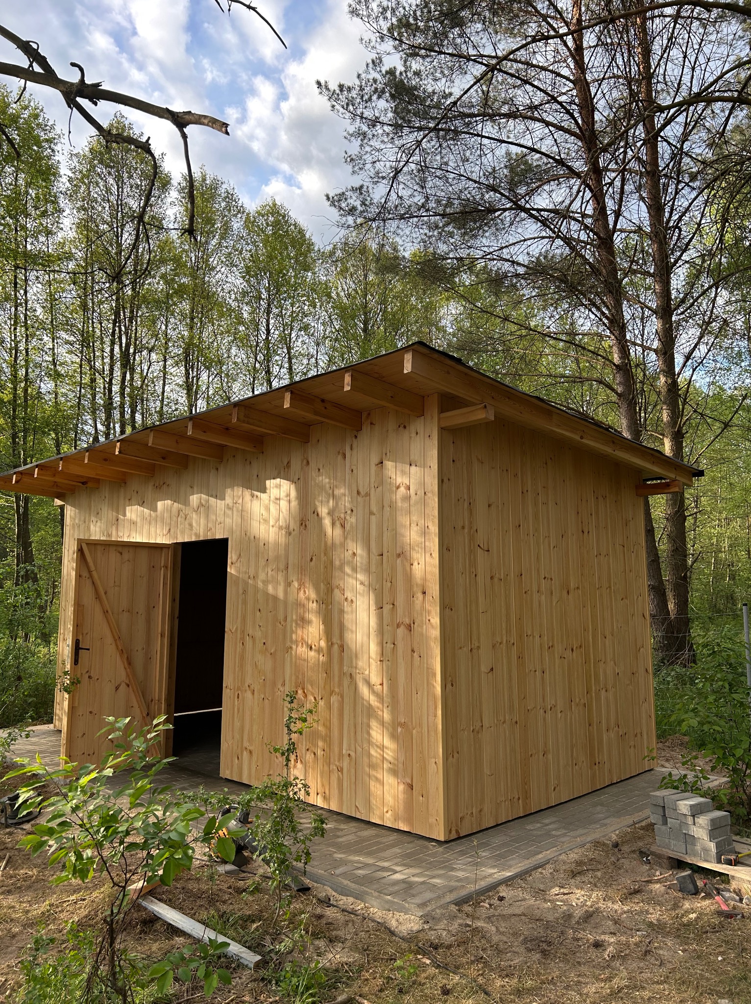 Nowy, drewniany domek narzędziowy z otwartymi drzwiami, usytuowany na obrzeżach lasu, na fundamencie z kostki brukowej. Widoczne narzędzia i materiały budowlane.