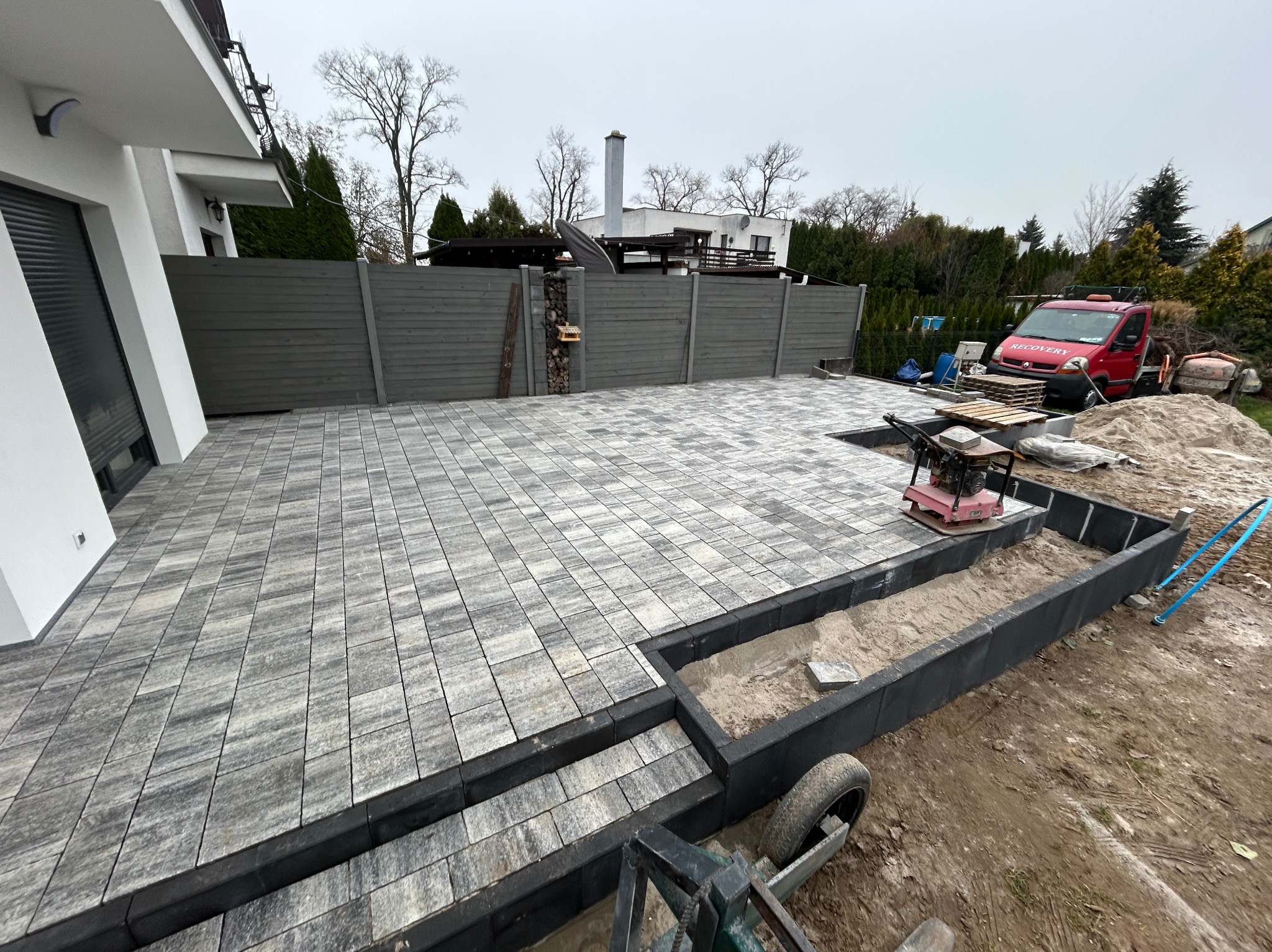 Ułożona kostka brukowa w ogrodzie, z fragmentem przygotowanym pod rabatę. W tle szary płot i budynek. Widoczny ubijak wibracyjny i auto dostawcze.