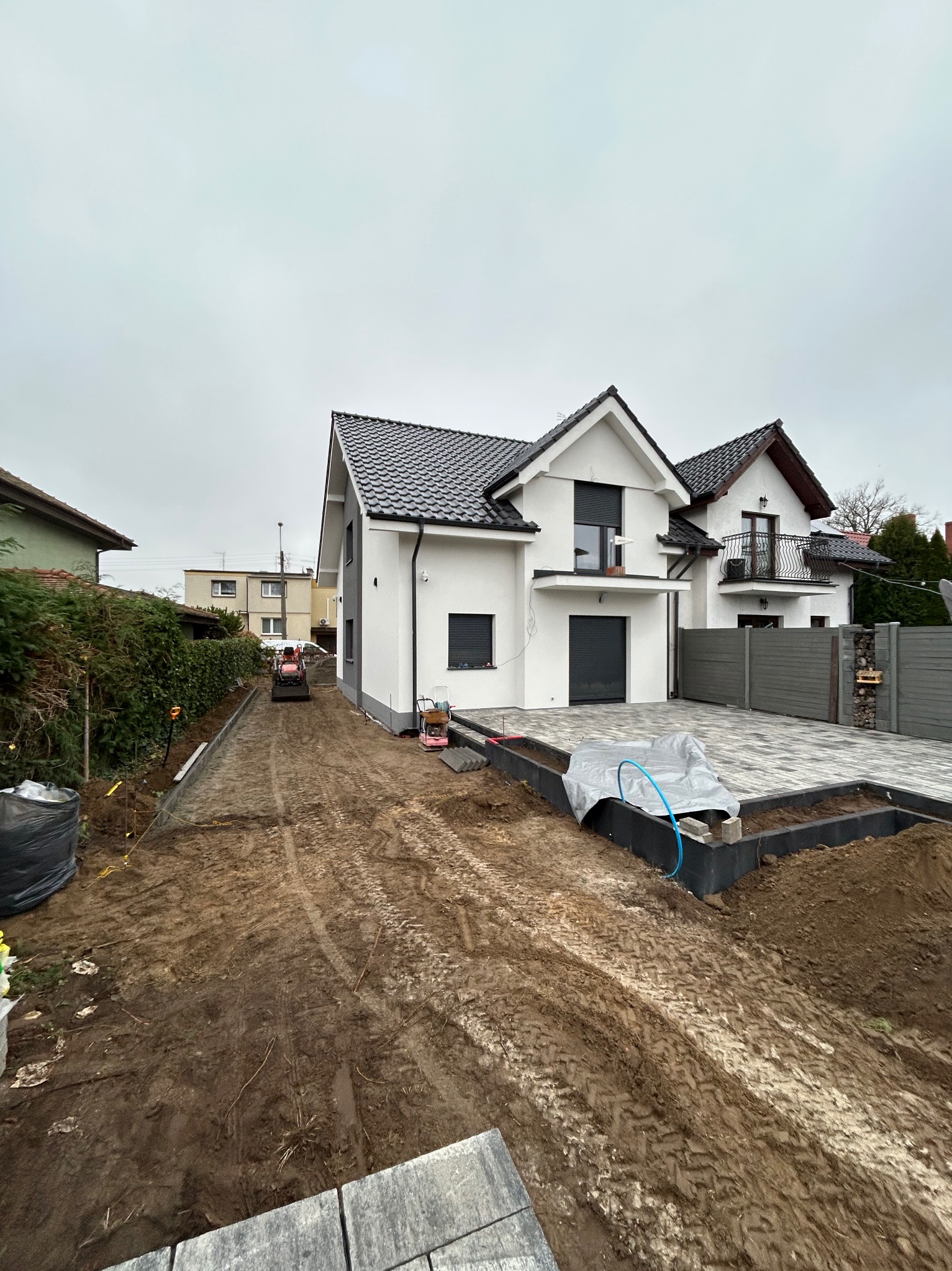 Nowoczesny dom jednorodzinny w trakcie budowy podjazdu z kostki brukowej. Widoczny minitraktor przygotowujący teren pod układanie kostki. Częściowo ułożona kostka.