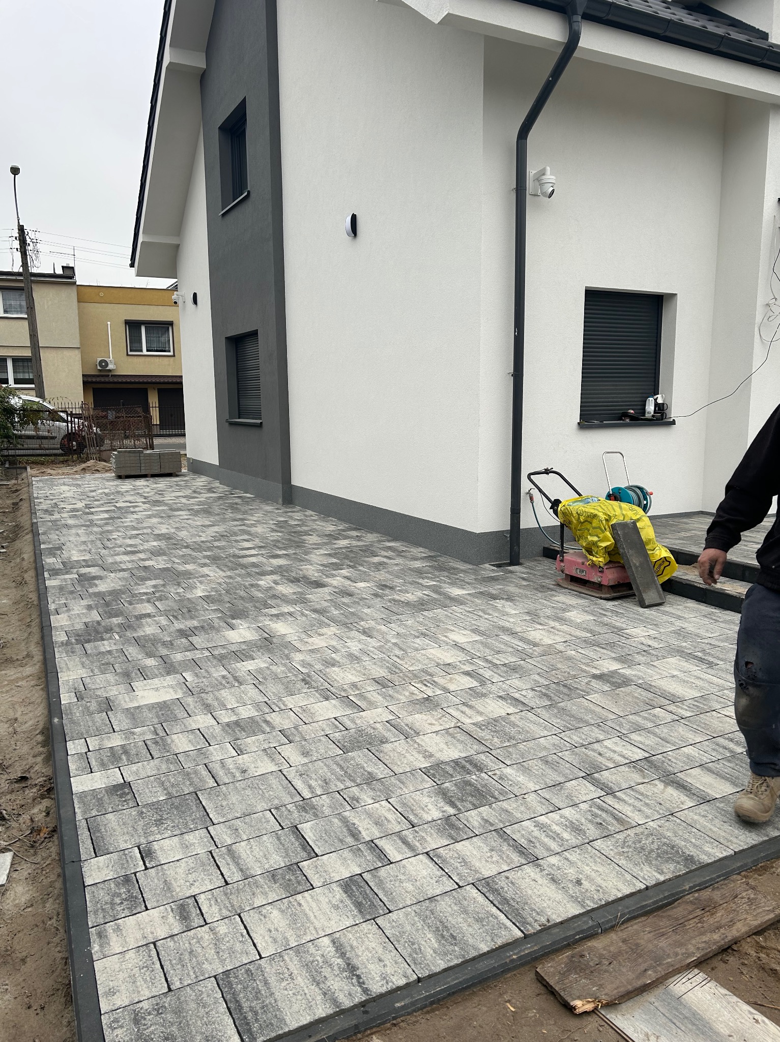 Ułożona szara kostka brukowa przed nowoczesnym domem. Widoczna ubijarka do kostki i fragment osoby. Kostka ułożona w nieregularny wzór.