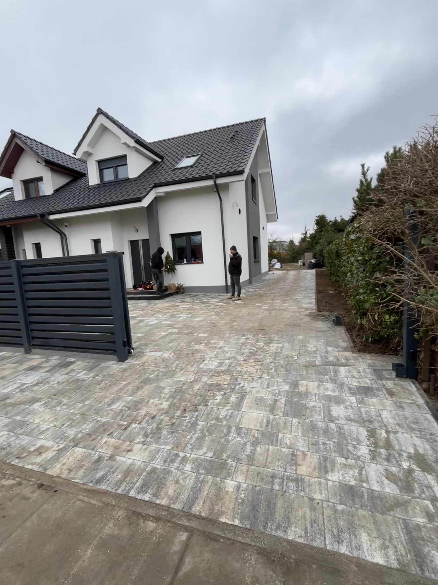 Nowoczesny dom z szarą, brukowaną aleją i ogrodzeniem z poziomych paneli. Dwóch mężczyzn stoi na posesji, jeden przy drzwiach, drugi na alei. Pochmurne niebo.