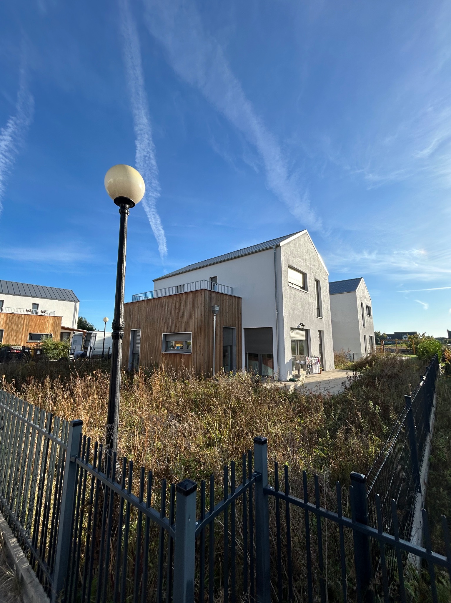 Nowoczesne domy z drewnianą elewacją i metalowym ogrodzeniem w słoneczny dzień. Charakterystyczna lampa uliczna. Błękitne niebo z smugami kondensacyjnymi.