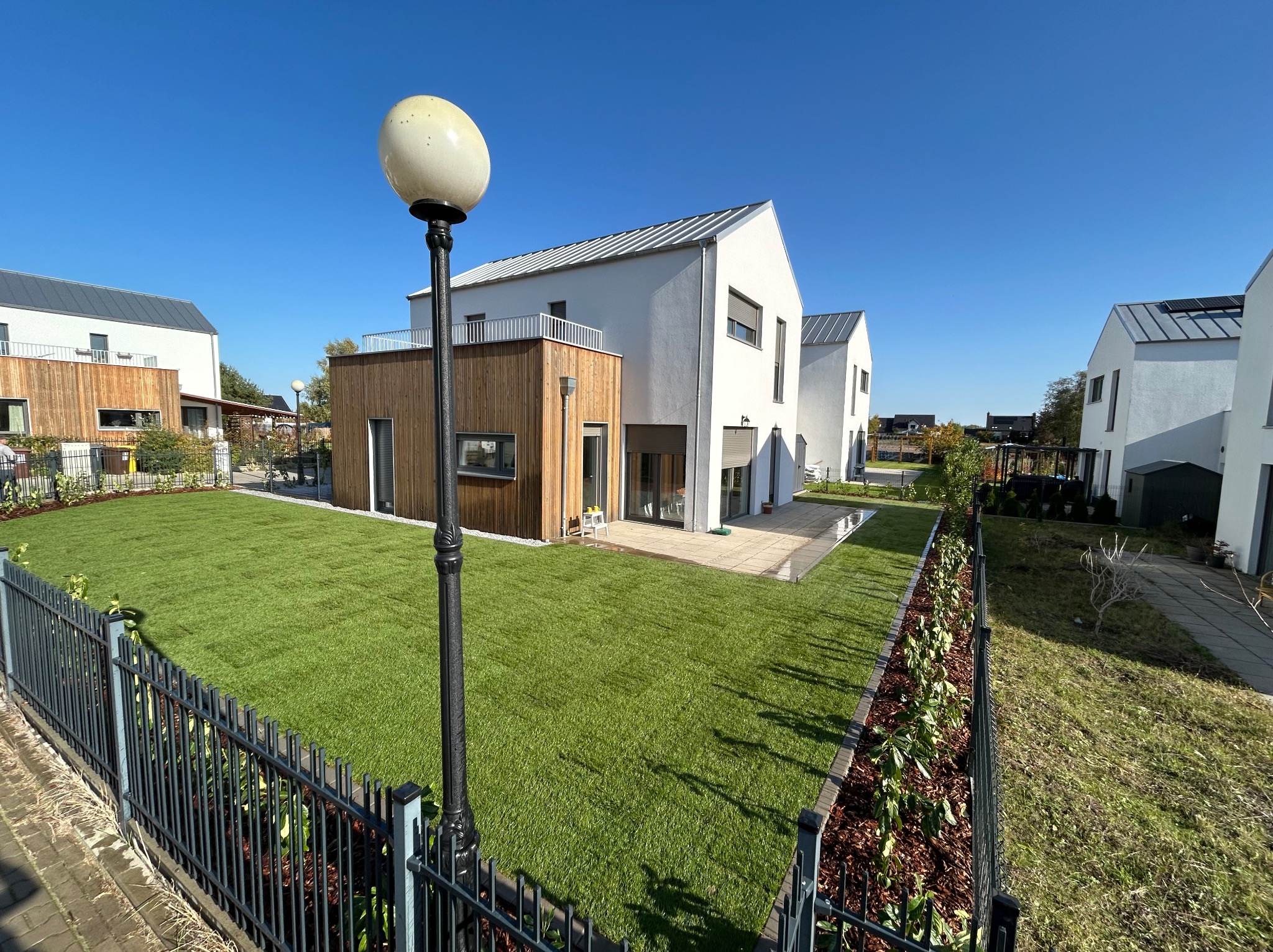 Nowoczesny ogród z równo ułożoną trawą z rolki, obramowany czarnym ogrodzeniem i rabatami z roślinami, na tle białych domów w słoneczny dzień.