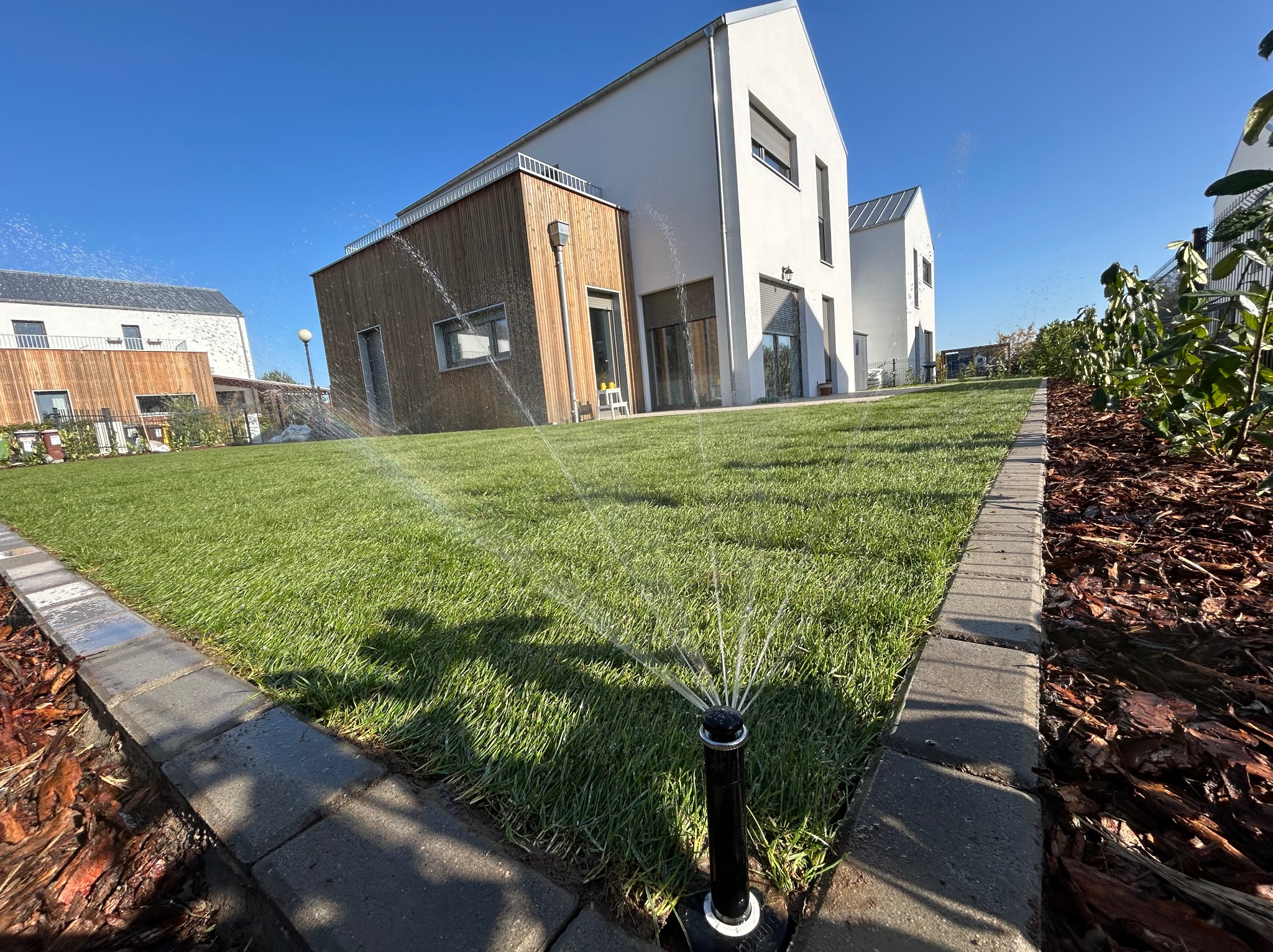 Automatyczny system nawadniania trawnika zraszaczami na tle nowoczesnego domu z drewnianą elewacją, w słoneczny dzień.