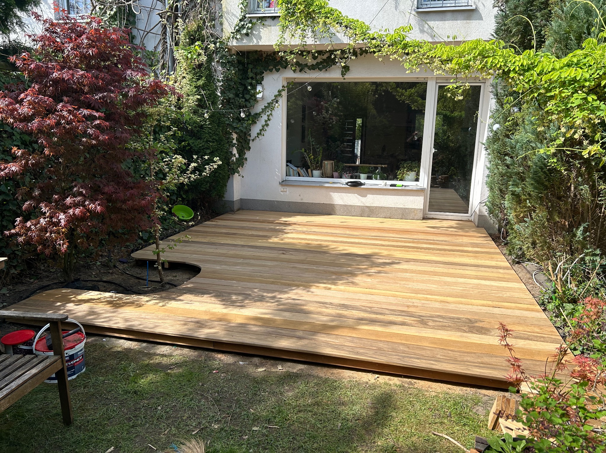 Nowy drewniany taras z jasnego drewna, częściowo ułożony, z wycięciem na drzewo, obok budynku z dużym oknem i zielenią. Widoczne farby i narzędzia.