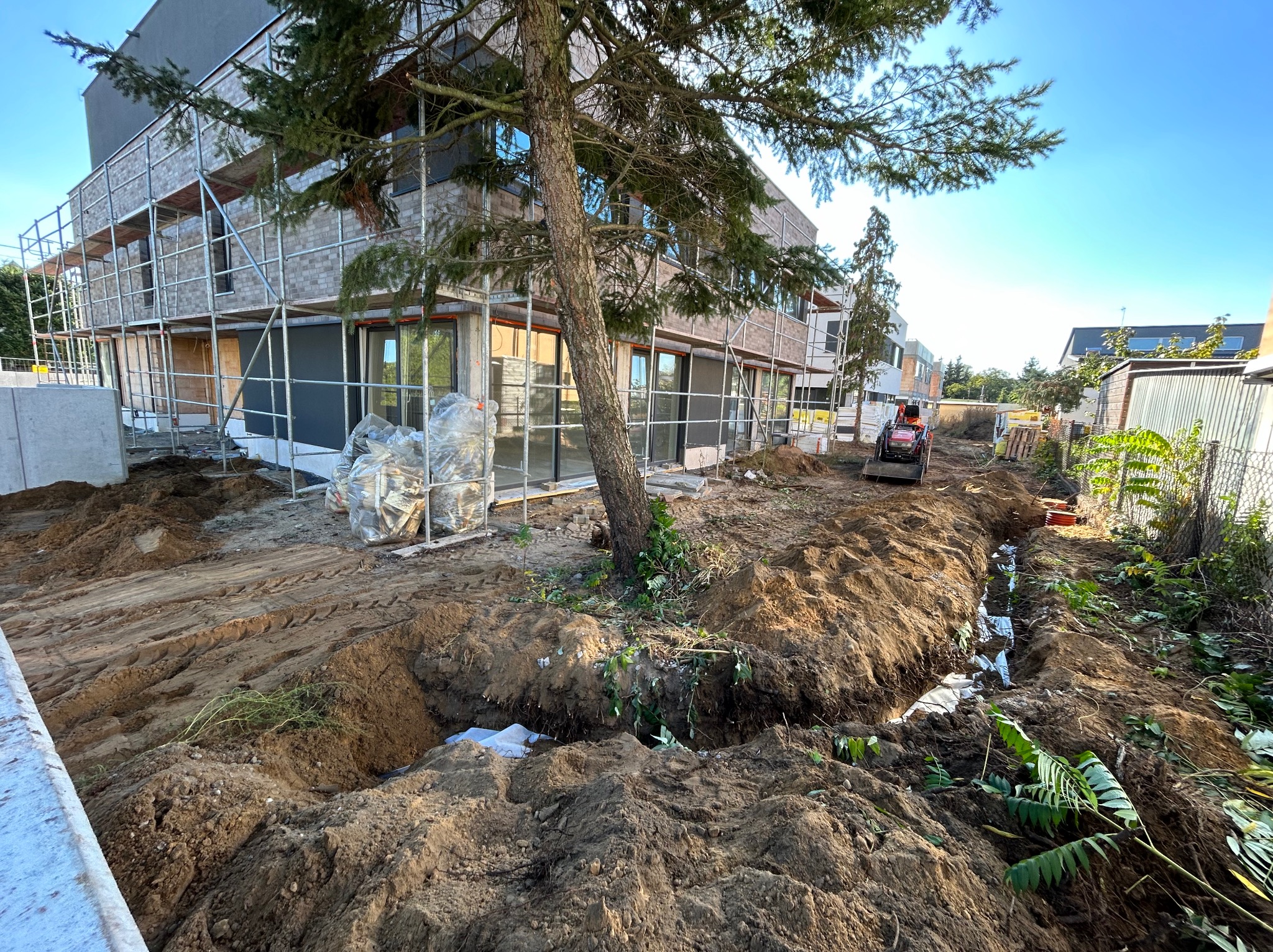Wykop pod instalacje przy nowoczesnym domu w trakcie budowy. Widoczny minikoparka i wykopany rów. Na pierwszym planie ziemia i gruz.