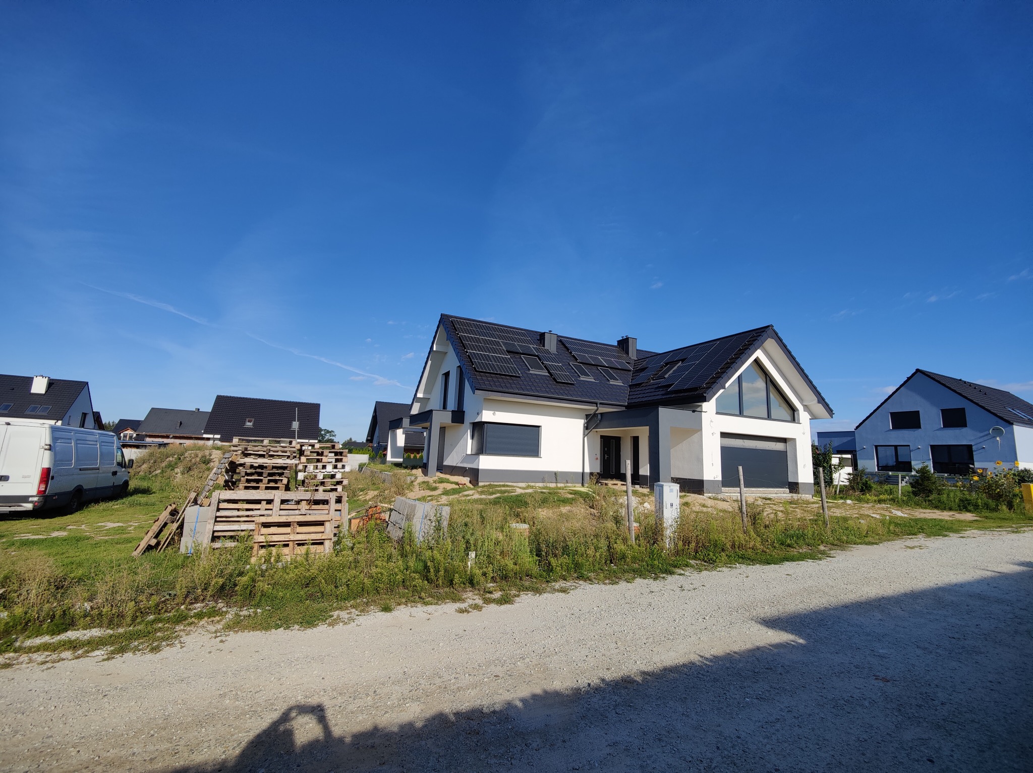 Nowoczesny dom jednorodzinny z panelami słonecznymi na dachu, widoczny z poziomu gruntu na tle błękitnego nieba, z elementami zagospodarowania terenu w trakcie realizacji.