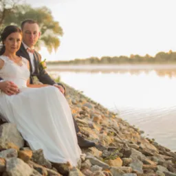 Para młoda siedząca na kamiennym brzegu rzeki w Szczecinie, w tle mgła nad wodą i drzewa w ciepłym świetle zachodzącego słońca.