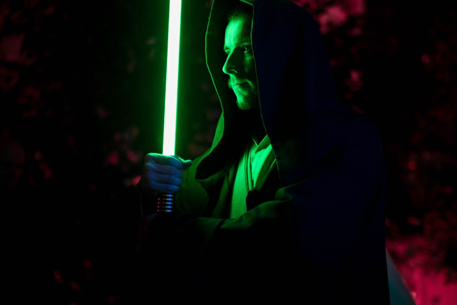 Mężczyzna w kapturze trzymający podświetlony na zielono miecz świetlny na ciemnym tle.