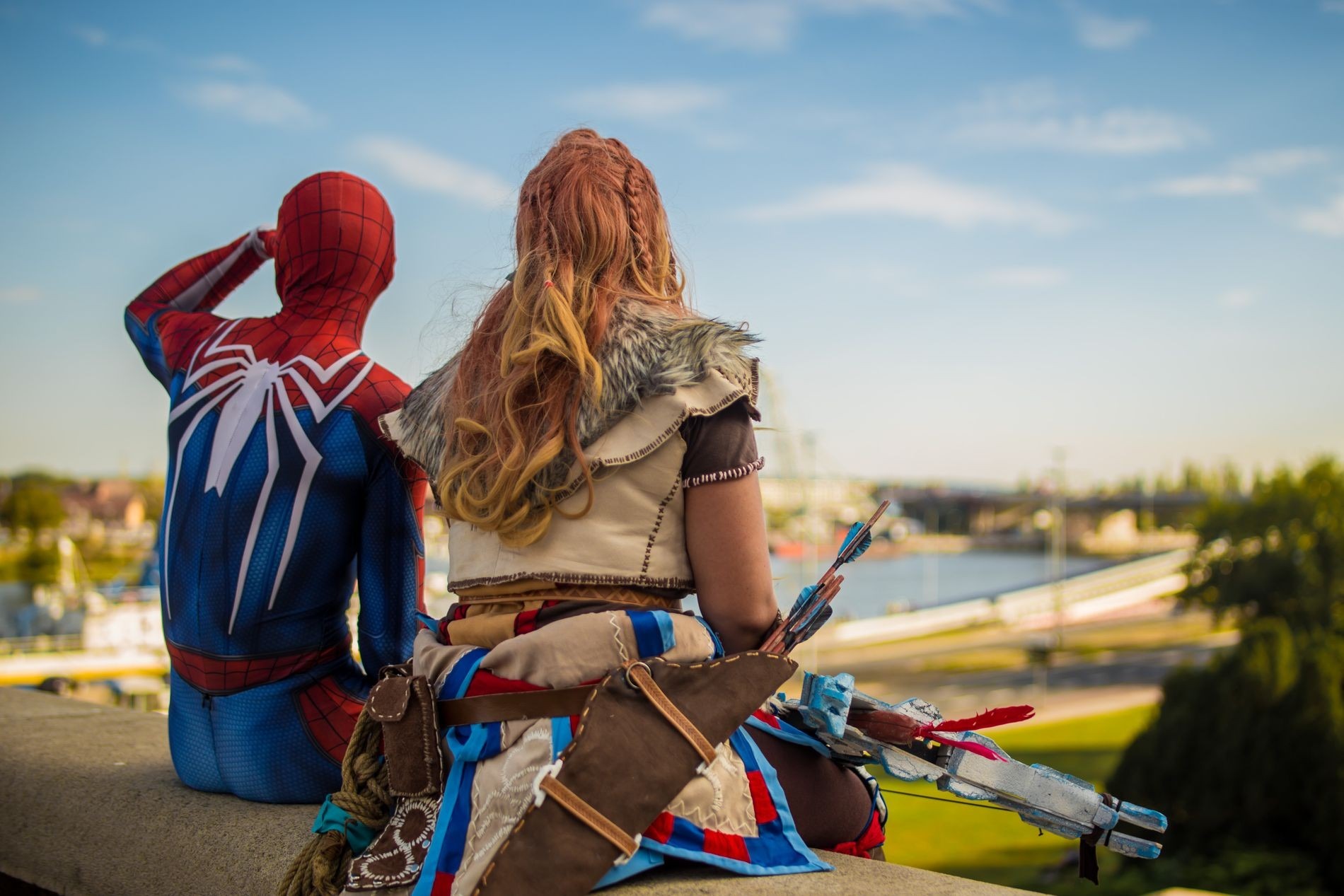 Dwie osoby w kostiumach: Spiderman w charakterystycznym czerwono-niebieskim stroju i kobieta przebrana za Aloy z gry Horizon Zero Dawn, siedzą na murku z widokiem na miasto i rzekę.