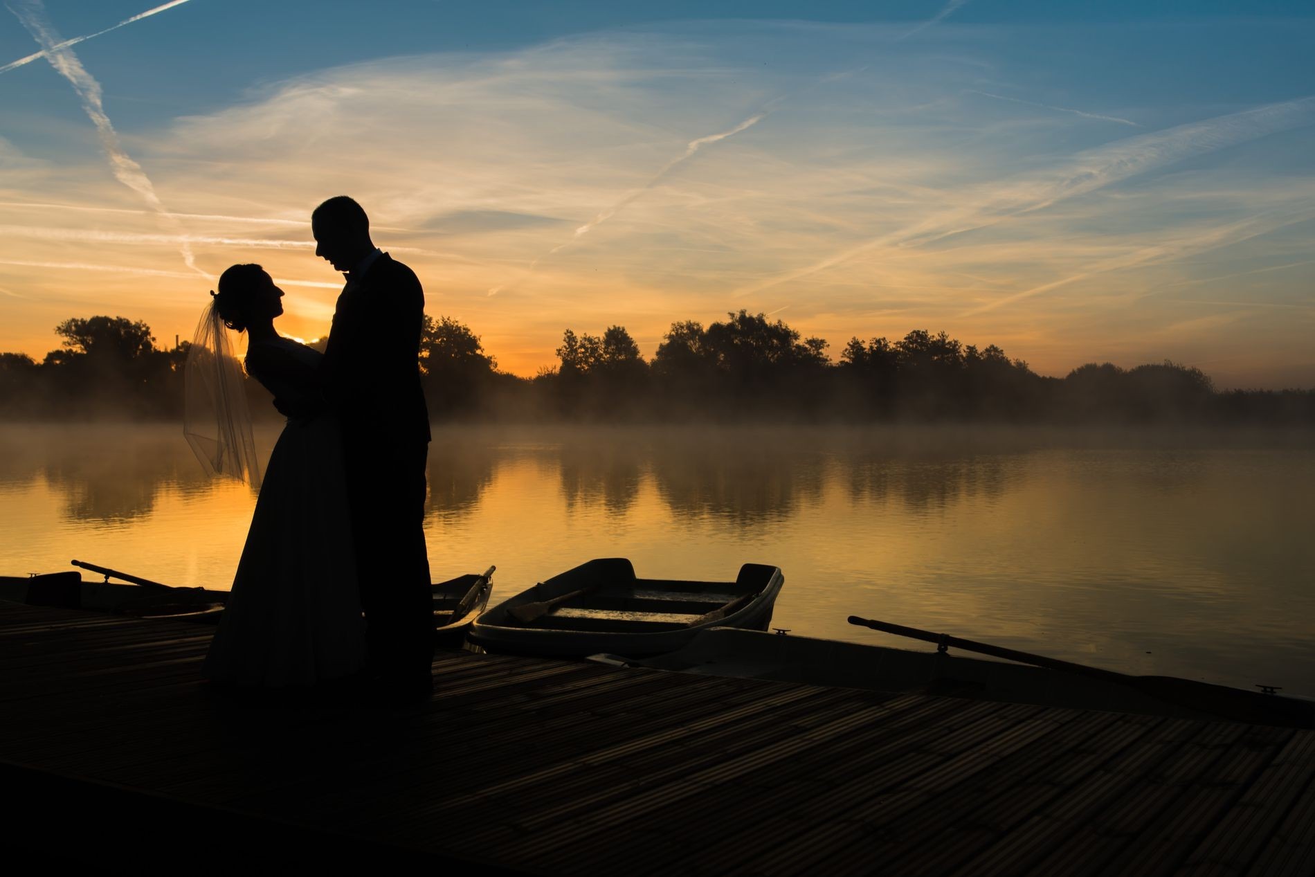 Silueta pary młodej na pomoście z łódkami, jezioro we mgle, zachód słońca i smugi kondensacyjne na niebie.