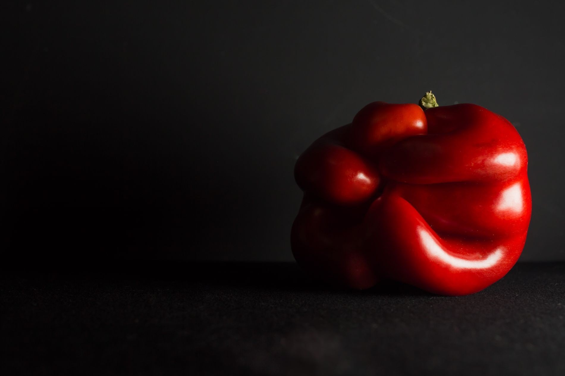 Czerwona papryka na ciemnym tle, oświetlona z góry, ukazująca jej nieregularny kształt i błyszczącą powierzchnię.