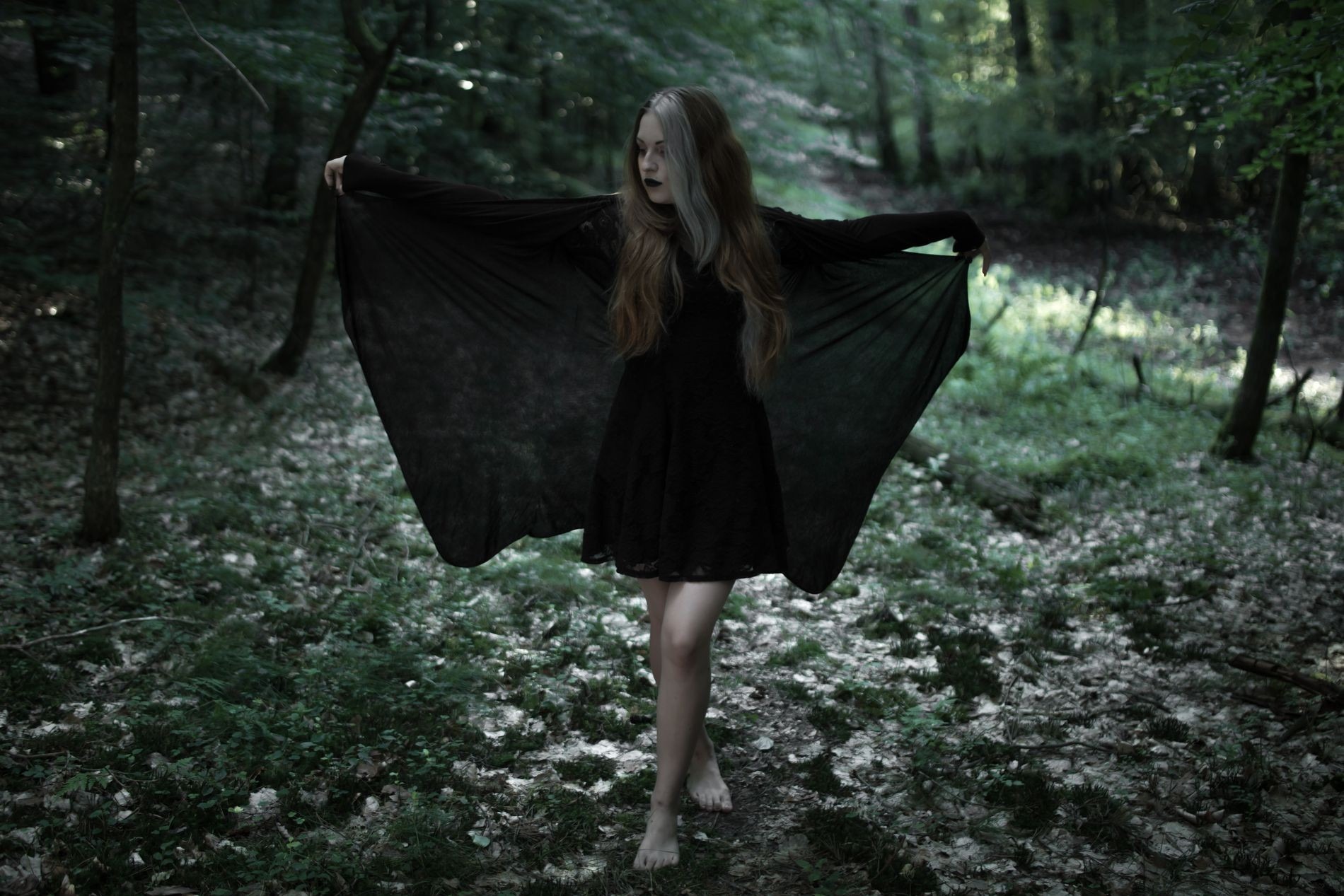 Kobieta w czarnej sukience i pelerynie pozuje boso na leśnej ścieżce, z dramatycznym makijażem i nietypową koloryzacją włosów.