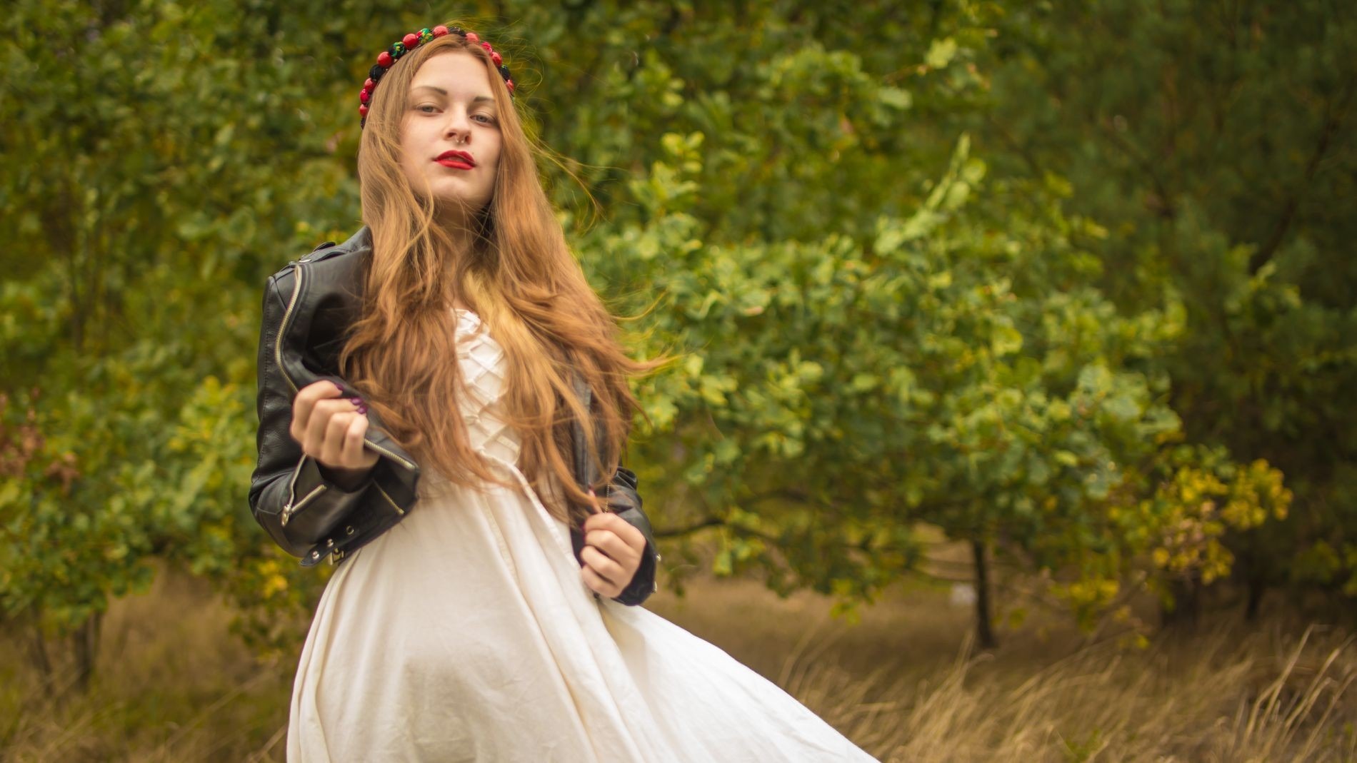 Rudowłosa kobieta w białej sukience i czarnej skórzanej kurtce, z wiankiem na głowie, pozuje na tle zieleni.