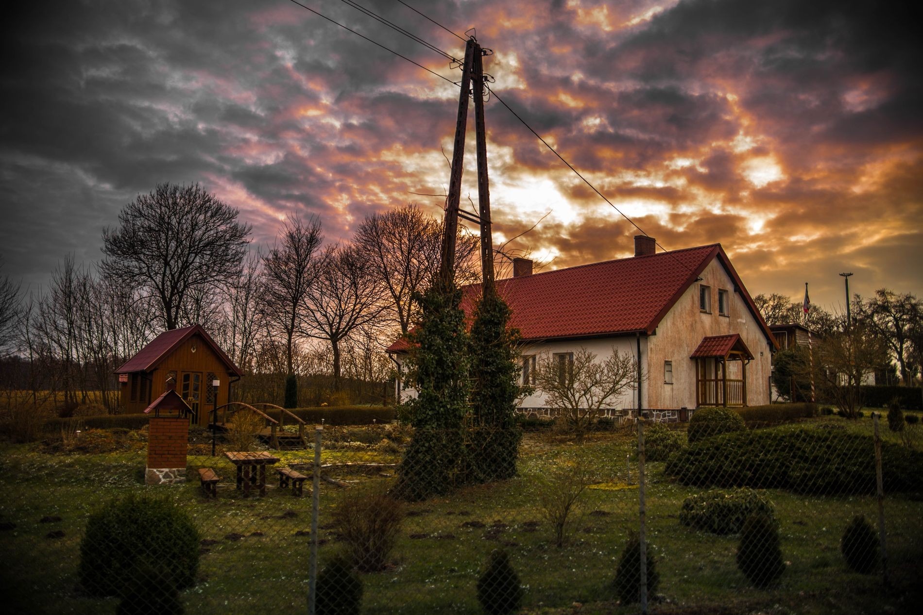 Wiejski krajobraz z domem, altaną i dramatycznym, pomarańczowo-szarym niebem o zachodzie słońca. Na pierwszym planie ogrodzenie z siatki.