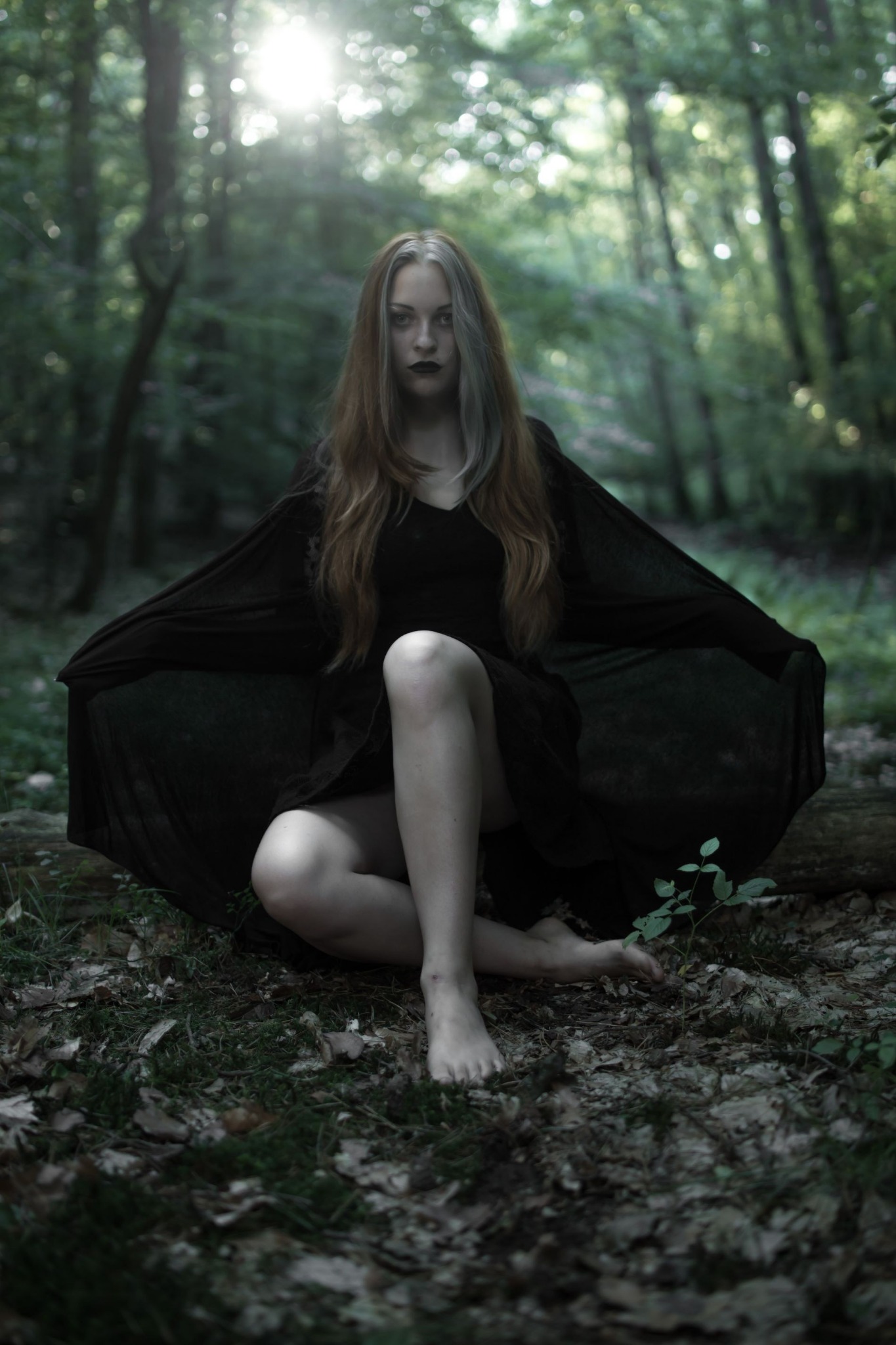 Kobieta w czarnej sukni pozuje boso w lesie, z bladą cerą i ciemnymi ustami, siedząc na ziemi z rozłożonymi rękawami sukni.