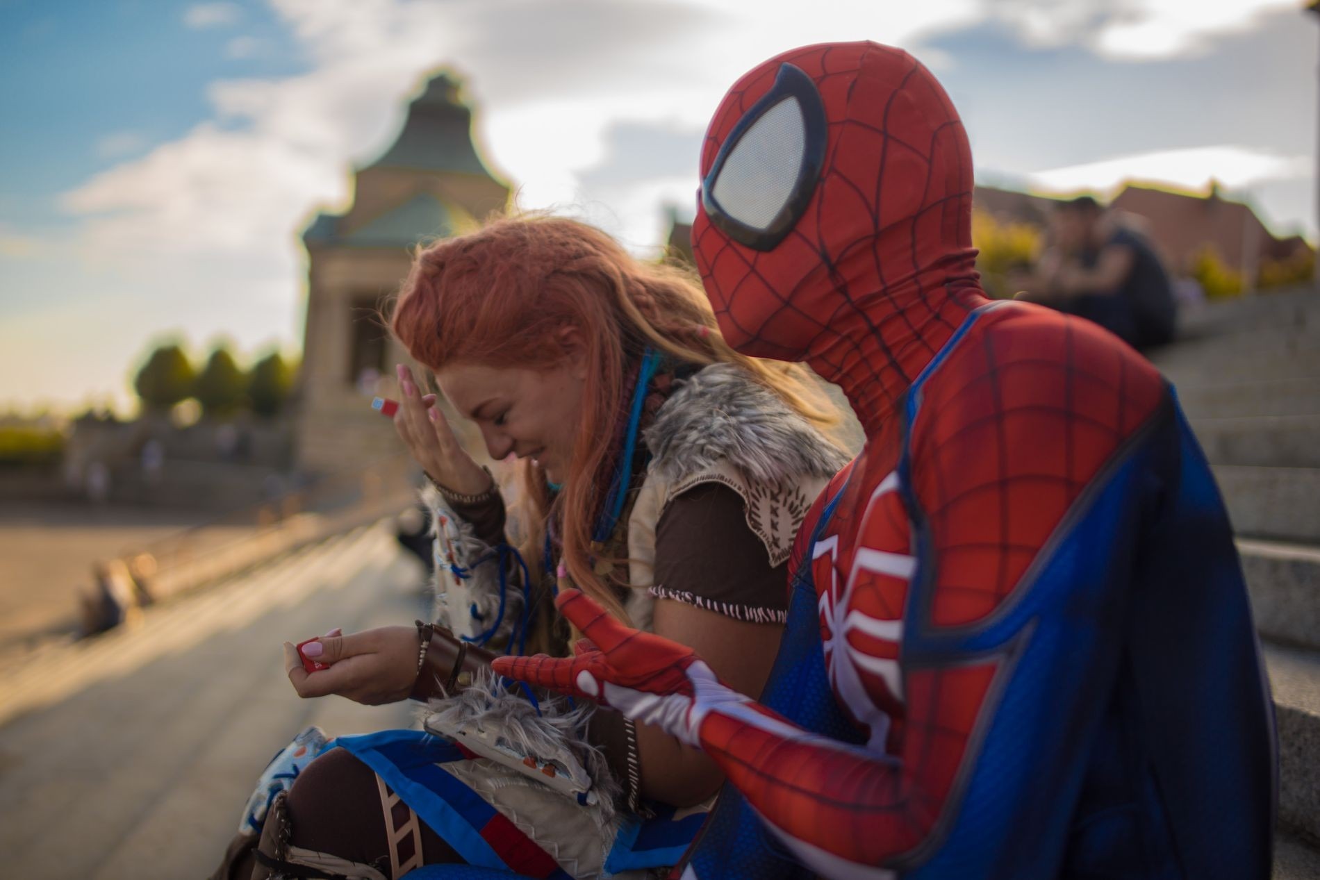 Kobieta przebrana za Aloy z Horizon Zero Dawn i Spider-Man siedzą na schodach, wymieniając się czerwonymi kapsułkami, z zabytkowym budynkiem w tle.