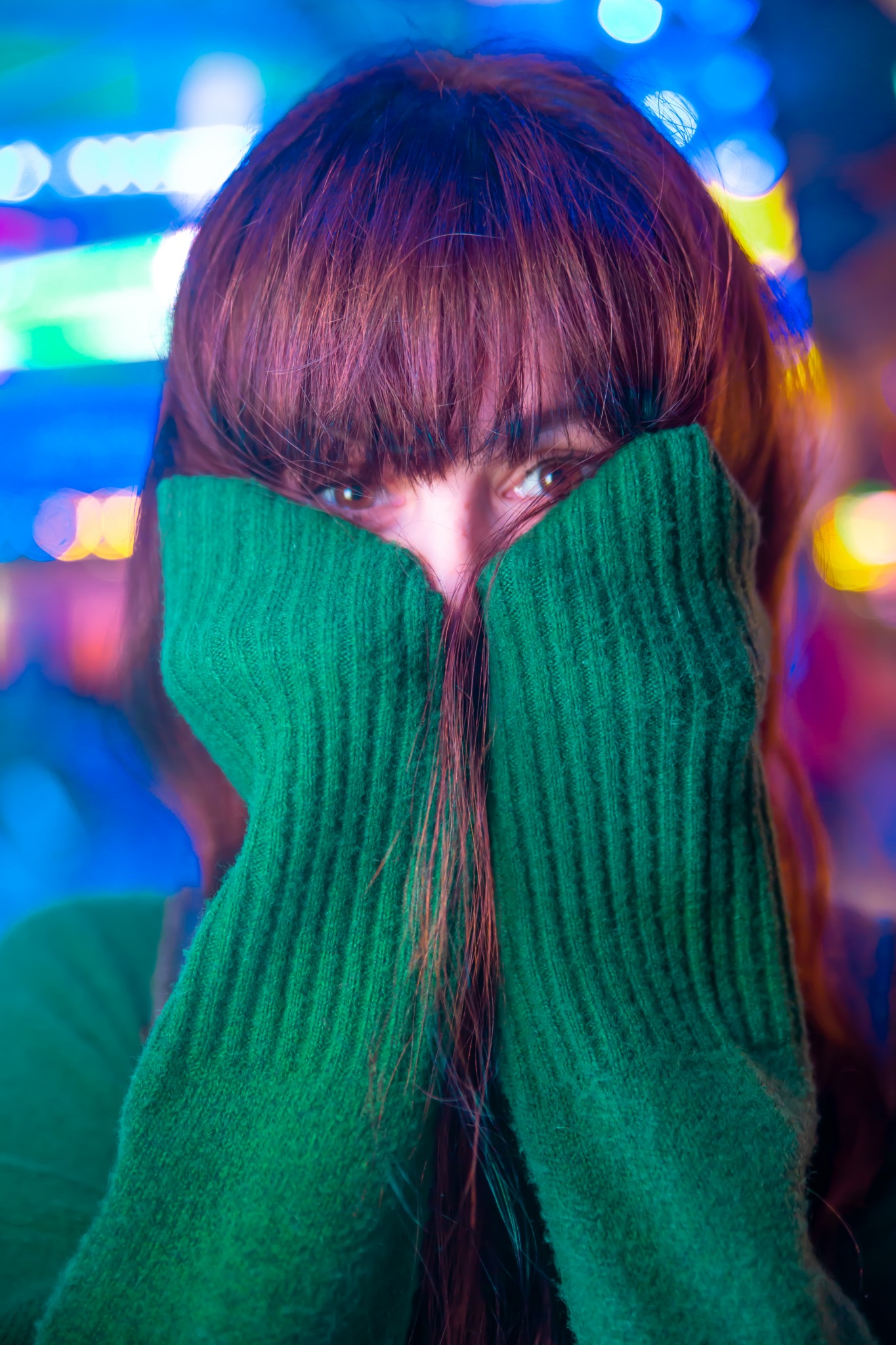 Portret kobiety zasłaniającej twarz zielonym, wełnianym swetrem na tle rozmytych świateł bokeh w odcieniach niebieskiego i żółtego.