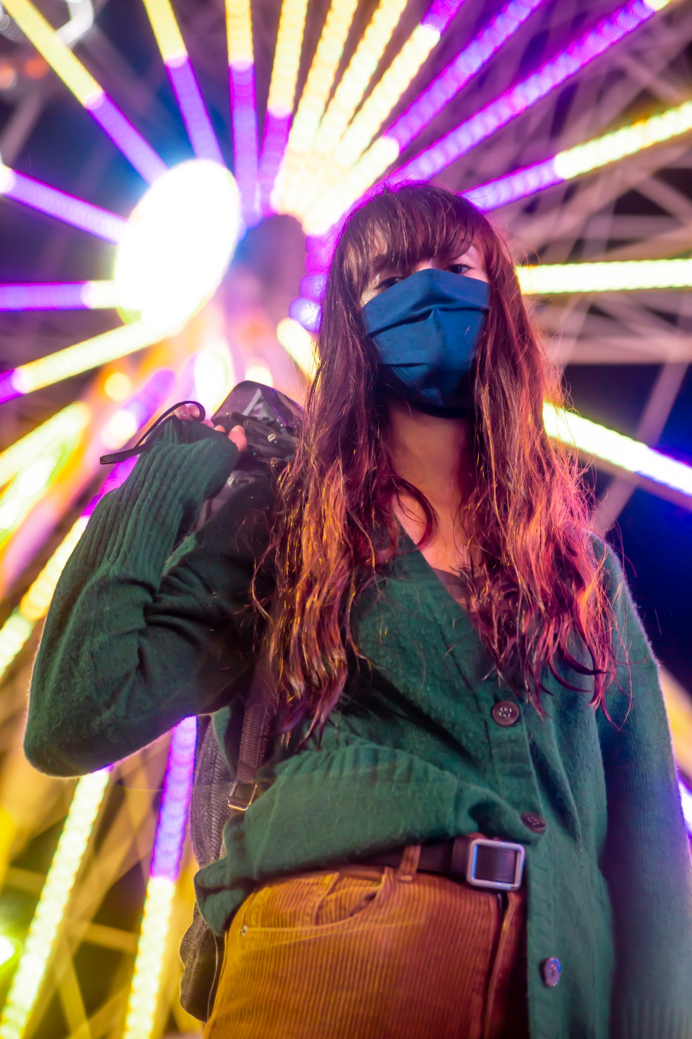 Kobieta w maseczce z aparatem na tle iluminowanego diabelskiego młyna, ujęcie z dołu.