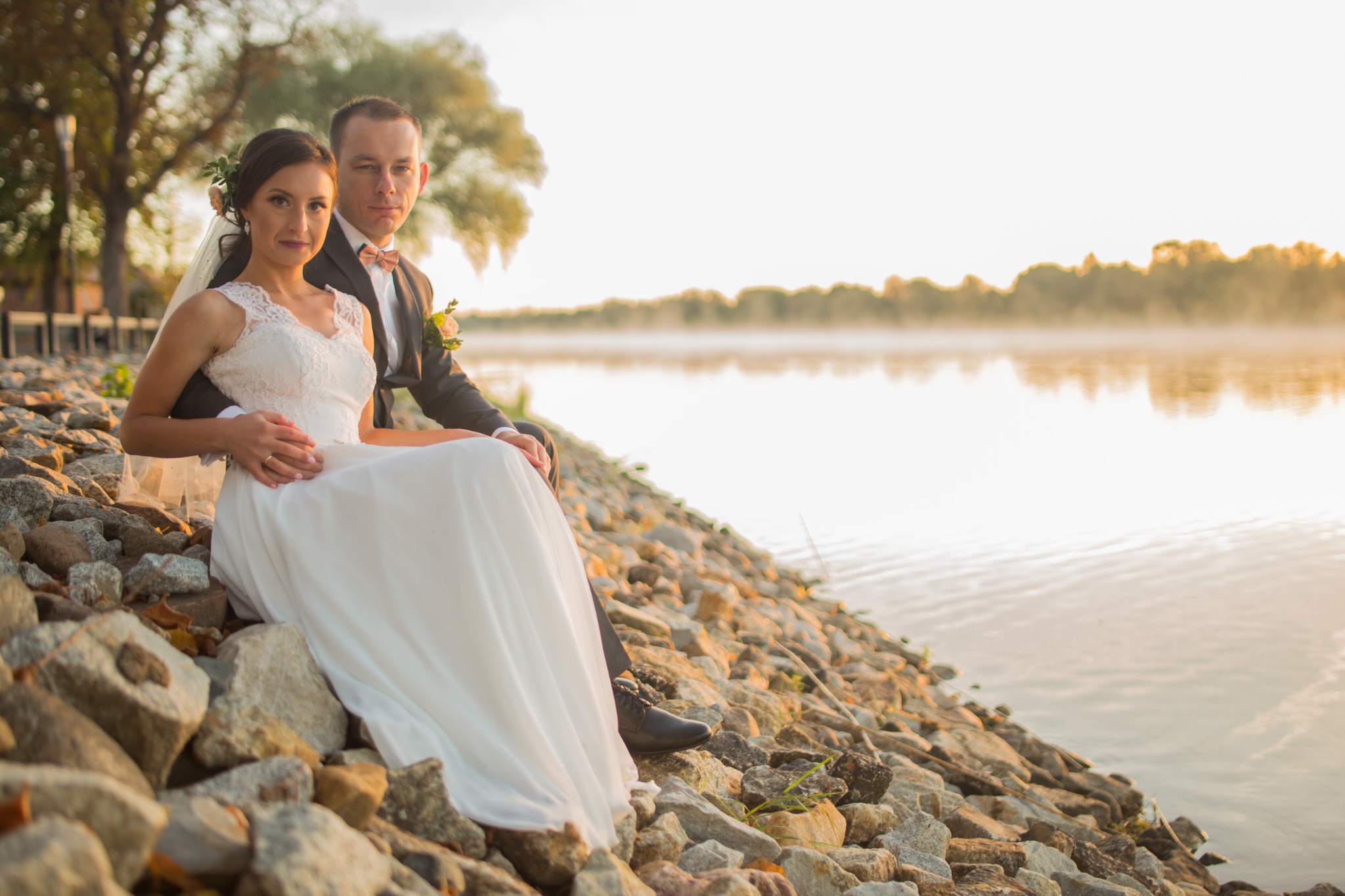 Para młoda siedząca na kamiennym brzegu rzeki w Szczecinie, w tle mgła nad wodą i drzewa w ciepłym świetle zachodzącego słońca.
