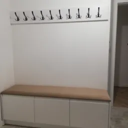 Wnętrze przedpokoju z białą szafką z tapicerowanym siedziskiem i wieszakiem z czarnymi haczykami na białej listwie.