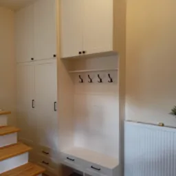 Biała zabudowa przedpokoju z szafami, siedziskiem i wieszakami, umieszczona przy drewnianych schodach