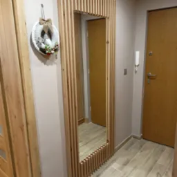 Nowoczesne lustro w drewnianej ramie z pionowych listewek, wiszący wianek świąteczny na ścianie, drewniane drzwi w tle, płytki podłogowe imitujące drewno.